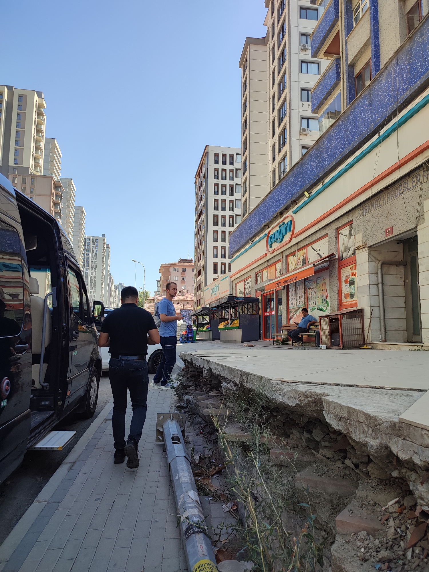 The image depicts a street scene in Kadıköy, Turkey. In the foreground, a damaged sidewalk with exposed bricks and dirt is visible. A person in a black shirt and jeans walks away from the camera on the adjacent paved walkway. To the left, the open door of a black van is visible. Further down the street, a man in a blue shirt stands on the sidewalk talking to another man. Behind them is a row of shops with awnings, and tall apartment buildings line the background. The sky is clear and blue.