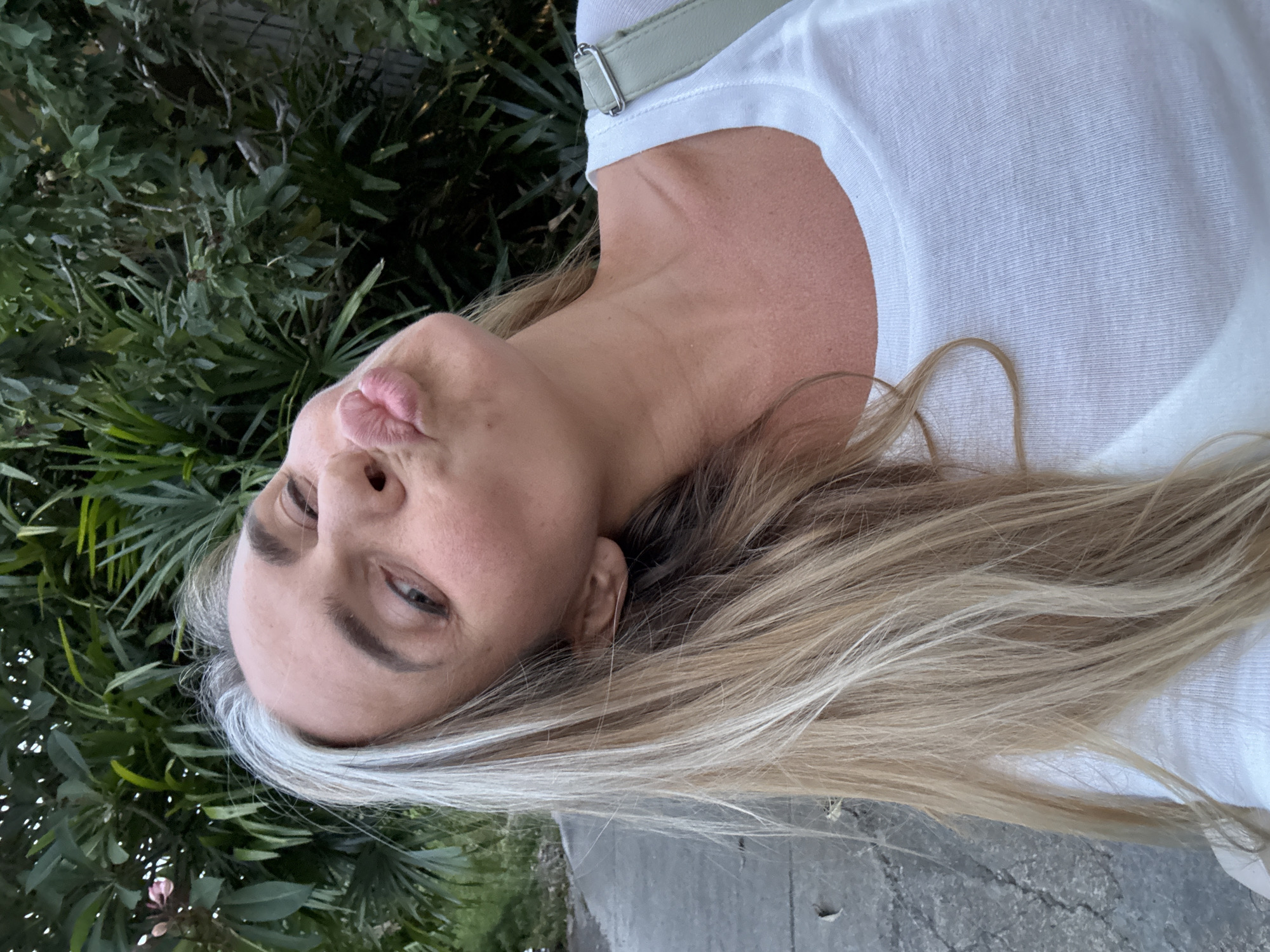 A woman with blonde hair and some gray strands is making a kissing face at the camera. She is wearing a white t-shirt. The background shows lush green foliage. The scene is set on a paved path next to the foliage. There are no explicit indicators to pinpoint the location within the city of Ko Samui District, Thailand.