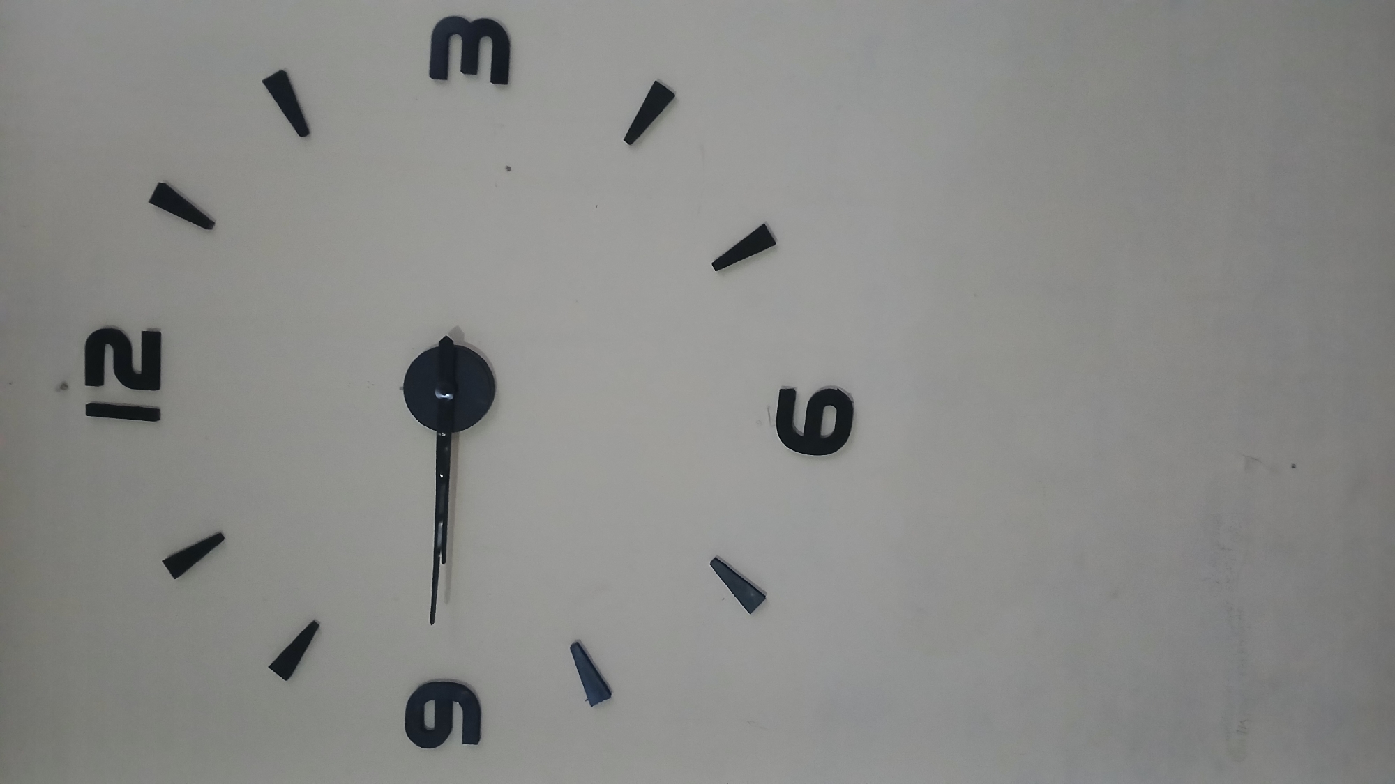 The image shows a close-up of a wall clock. The clock has a minimalist design with black numbers and black hands. The numbers 12, 3, 6, and 9 are visible. The hour hand points to the 9, and the minute hand points to the 12. The clock appears to be a modern, decorative piece. There is no indication of its location in East Jakarta, Indonesia, or any other specific geographic area. The background is a plain, light-colored wall, possibly painted or covered in a neutral material. There are no people, actions, or interactions depicted in the image. The environment is static and focused solely on the clock.