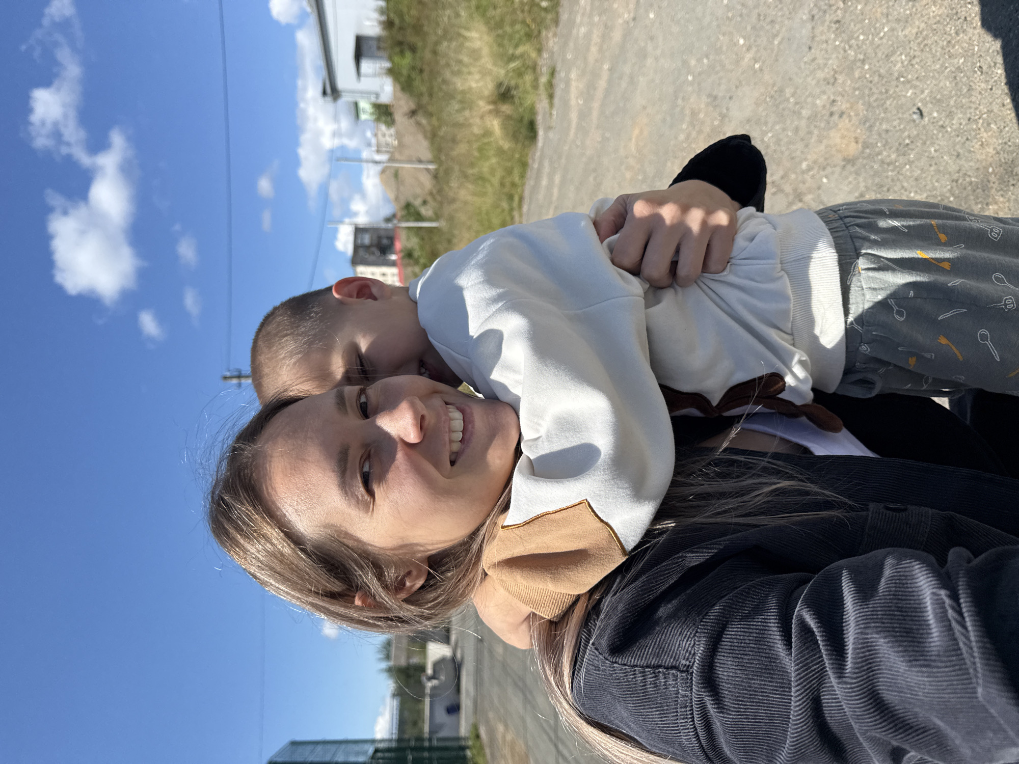 A medium close-up selfie shows a woman and a young child embracing outdoors on a sunny day with a bright blue sky and scattered white clouds. The woman, with long blonde hair and fair skin, smiles at the camera. She wears a dark, ribbed jacket over a white top. The child, facing away from the camera and partially obscuring the woman's face, is hugged tightly by the woman. The child wears a white sweatshirt with tan accents on the sleeves and gray sweatpants patterned with white and orange utensils. They are standing on a gravel path in what appears to be an industrial or semi-developed area. In the background, there are industrial buildings, construction materials, and grassy areas. The location is identified as Gornozavodsk, Russia.