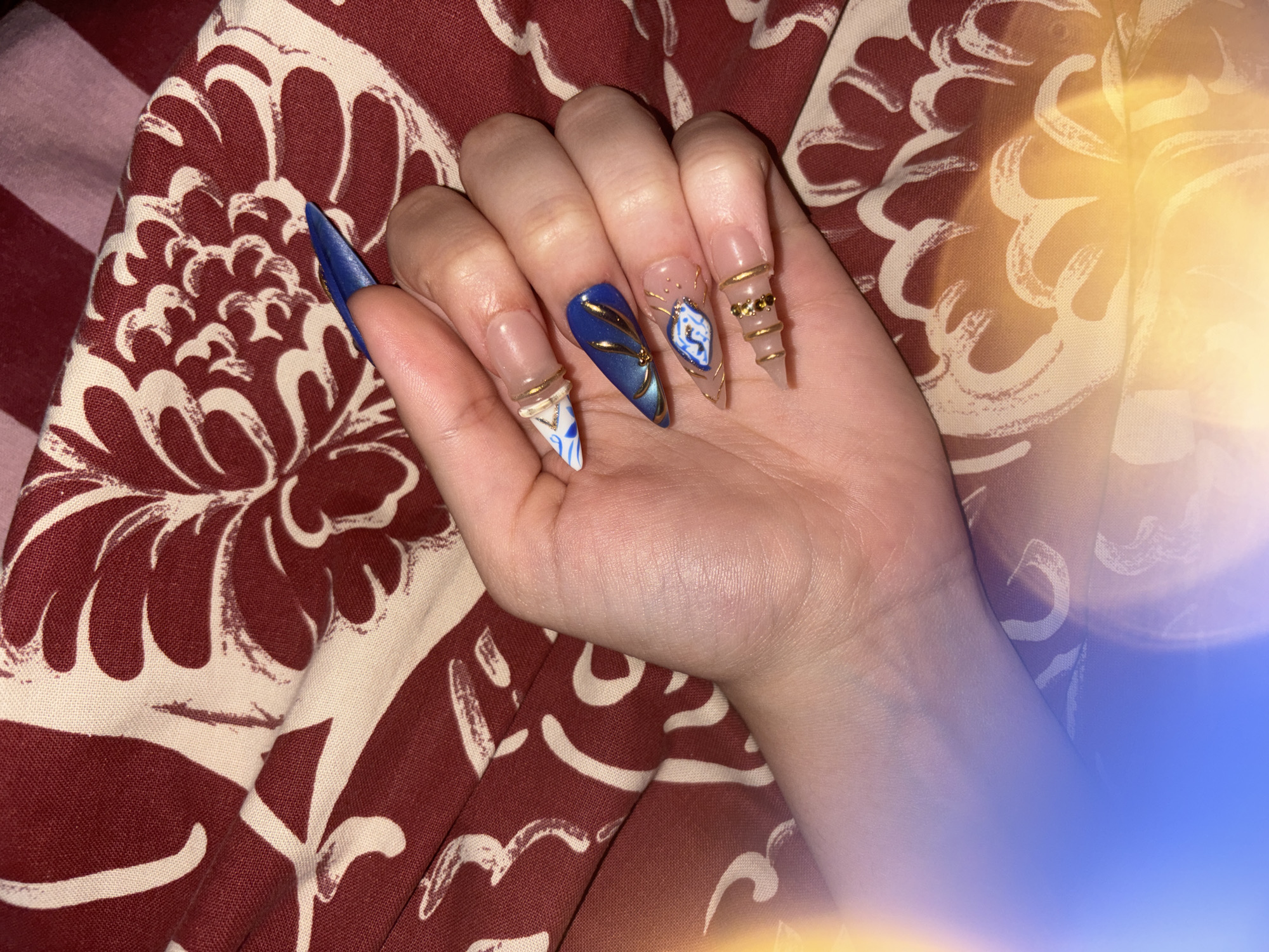 The image displays a close-up of a person's left hand, palm up, showcasing an elaborate set of long, decorated artificial nails. The hand rests on a reddish-brown fabric patterned with cream or off-white floral or swirling designs.
Each visible nail features a distinct design. The pinky nail is clear and pointed, adorned with multiple gold rings wrapped around it, some featuring tiny gems. The ring finger nail is also clear and pointed, embellished with a blue and white patterned design, possibly resembling a fragment of pottery or a stylized emblem, outlined by thin gold lines and dots. The middle finger nail is a solid deep blue with a pointed shape, accented with metallic gold lines or a reflective sheen. The index finger nail is clear and pointed, sporting a blue and white geometric or abstract pattern, complemented by a gold ring near its base. The thumb nail, partially visible, is blue and pointed, similar in color to the middle finger.
The hand appears youthful and well-manicured. The lighting suggests an indoor setting, with noticeable lens flares in yellow-orange and blue hues appearing on the right side of the image, creating a soft, artistic glow. There is no visible text in the image. The scene emphasizes the intricate and diverse nail art.