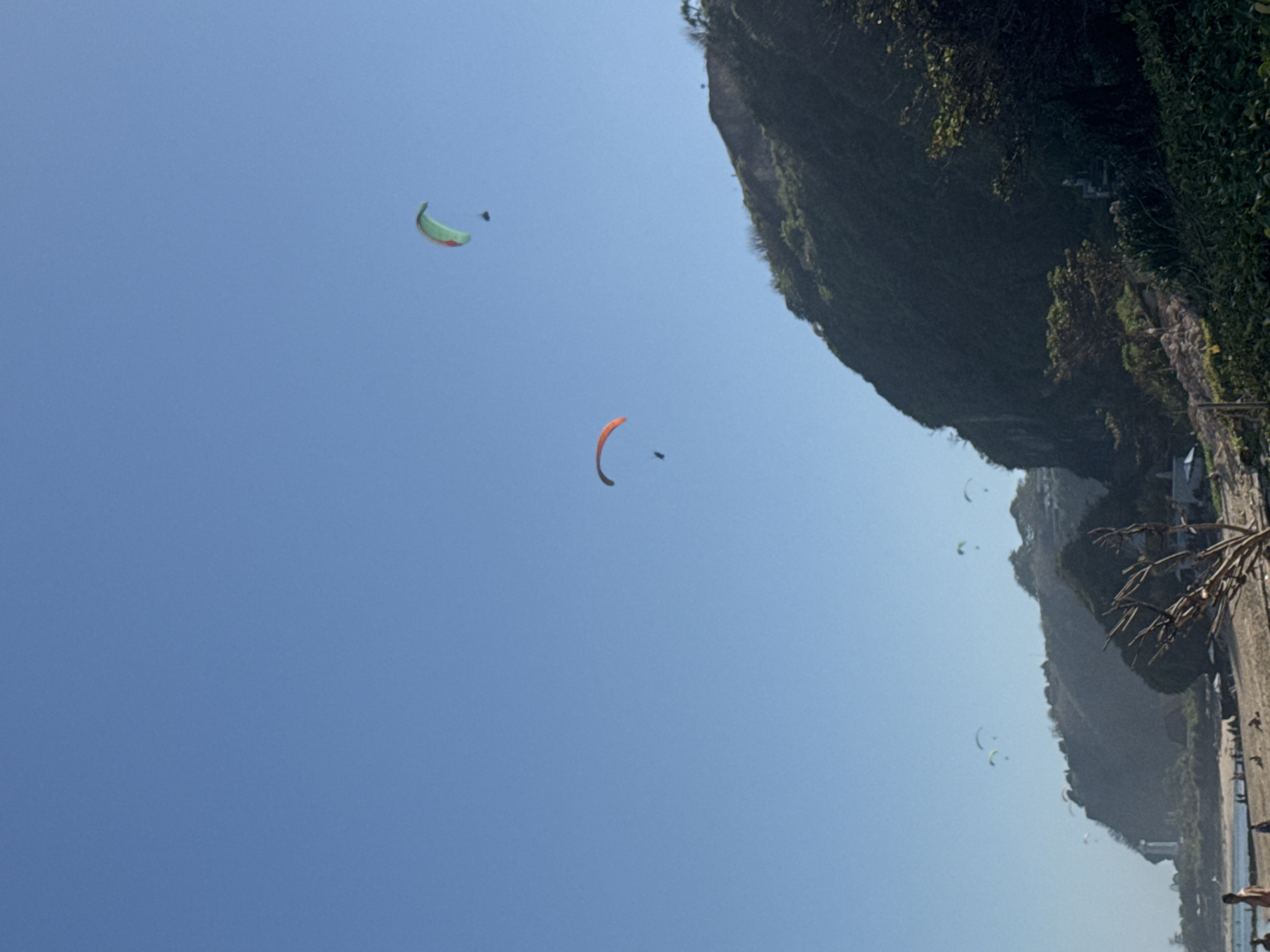 The image shows a clear blue sky over Kuta Selatan, Indonesia, with a beach and a dramatic cliff face in the foreground. Several paragliders are visible in the sky, enjoying the coastal scenery. The beach appears to be populated with a few people, and the cliff face is covered in lush greenery. A lighthouse is visible in the distance on the left side of the image. The scene evokes a sense of leisure and adventure, with the paragliders adding a dynamic element to the otherwise tranquil beach landscape.