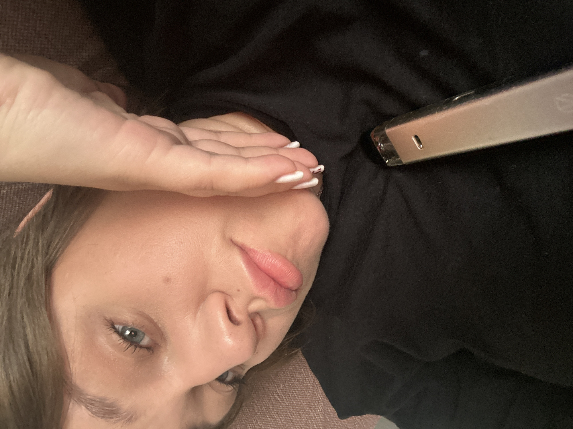 An overhead shot shows a person lying down, looking directly at the camera. The subject appears to be a young adult with light skin, light brown hair, and blue eyes. Their right hand is raised, partially covering their forehead and the upper part of their face, with fingers slightly spread. The visible nails are painted with a white tip, resembling a French manicure, and one finger has a small dark detail.
The person is wearing a black top or shirt, which covers most of their body visible in the frame. Their lips are full, with a natural pink hue, and their gaze is steady towards the viewer. Next to their head, on the black fabric, rests a slim, metallic object. This object is light-colored, possibly silver or champagne, and features a small rectangular port or indicator light on its side. It resembles an electronic device, such as a vape pen. A very faint, unidentifiable logo or text is barely discernible on the object's surface.
The background suggests an indoor setting, possibly a couch or bed, with a textured, light brown or greyish upholstered surface visible on the left side and in the lower portion of the image. The overall lighting is soft, creating a calm and intimate atmosphere. There are no explicit location cues within the image to indicate Chaikovsky, Russia.