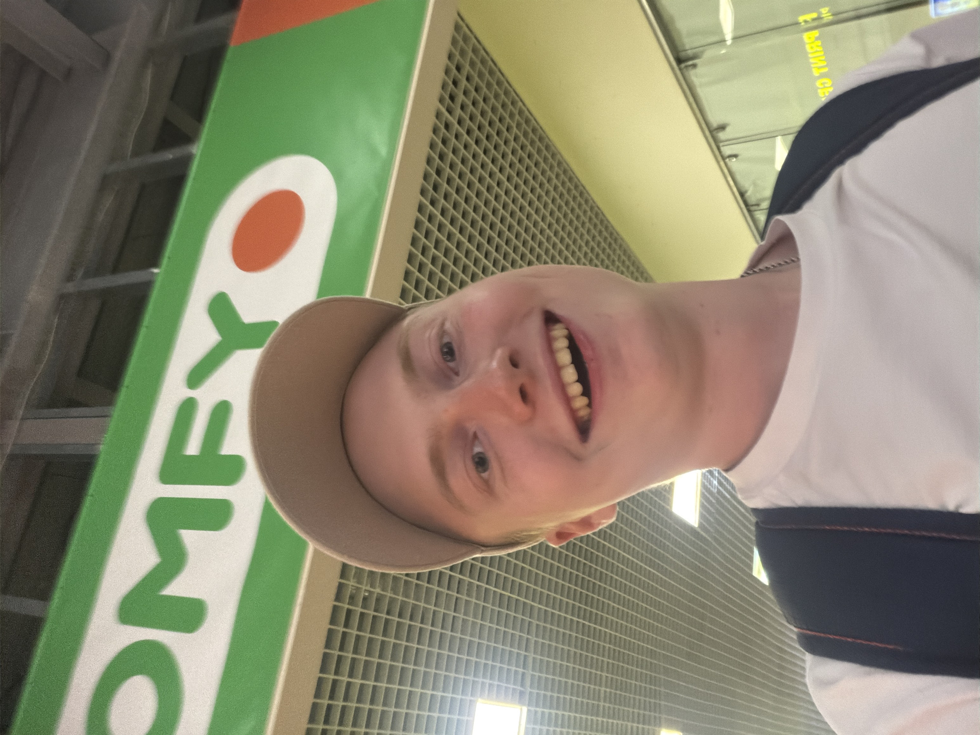 A young man with fair skin, light brown hair, and a light brown baseball cap is taking a selfie. He is smiling widely, showing his teeth. He is wearing a white t-shirt and a dark backpack. Behind him is a sign with green and white lettering that says "COMFY" with a red oval. The ceiling above him is made of metal grids with fluorescent lights. In the background, there are glass windows with some text visible, which appears to be in Ukrainian. The overall impression is that of an indoor environment, possibly a shopping mall or an electronics store.
