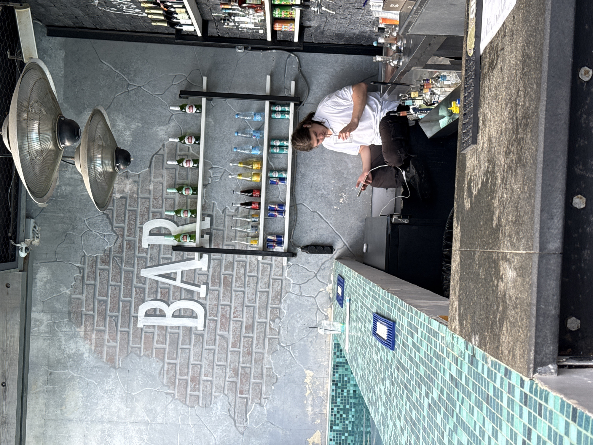The image depicts a bar area, possibly in Yalta, Ukraine, characterized by a modern, industrial aesthetic. The wall behind the bar features a distressed concrete finish with an inset brick pattern and large white "BAR" lettering. Two large, industrial-style heaters hang from the ceiling. Shelves on the wall display an assortment of bottles, including beer and what appear to be spirits, along with cans of Red Bull. In the foreground, a bar counter with a turquoise mosaic tile finish runs along the edge of what seems to be a pool. A young man with long brown hair, wearing a white polo shirt and dark pants, is seated on a stool behind the bar. He is holding a smartphone in his left hand and a glass with a straw in his right, appearing to drink from it. He has earbuds in his ears. The bar itself is equipped with bottles, glassware, and what looks like a beer tap system. The overall atmosphere is casual and contemporary.