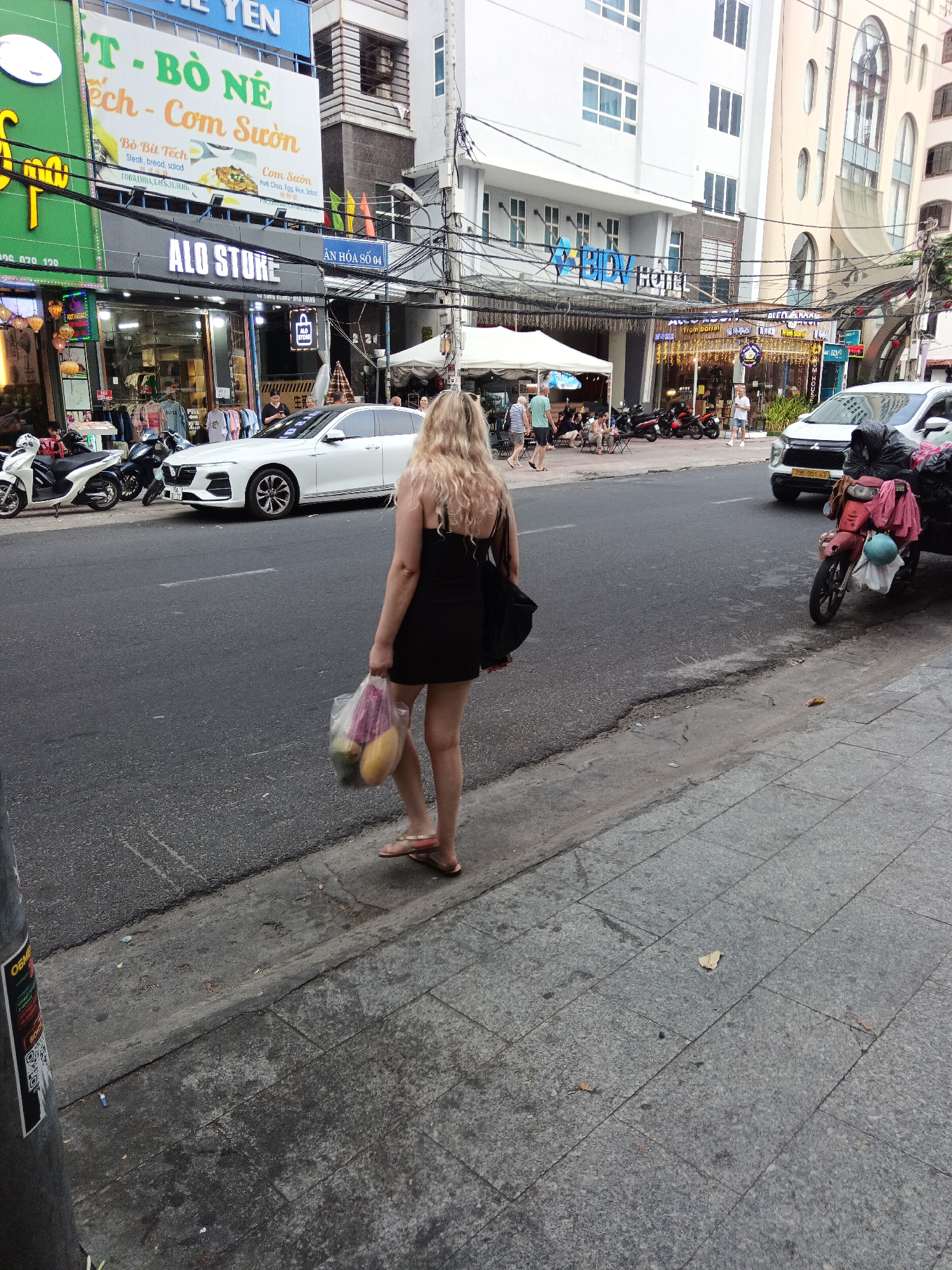 The image depicts a street scene in Nha Trang Ward, Vietnam. A woman with blond hair, wearing a short black dress and flip-flops, stands on a sidewalk, holding a plastic bag filled with what appear to be fruits. She faces away from the camera, towards a street with passing vehicles and parked motorbikes. Behind her are various businesses, including one called "ALO STORE" and a restaurant with the sign "Bò Bít Tếch". In the background, there's a building marked "BIDV HOTEL". Power lines are visible overhead, crisscrossing the scene. The sidewalk is tiled and slightly worn, with some debris scattered on the road. A utility pole with stickers attached to it is visible on the left edge of the frame.