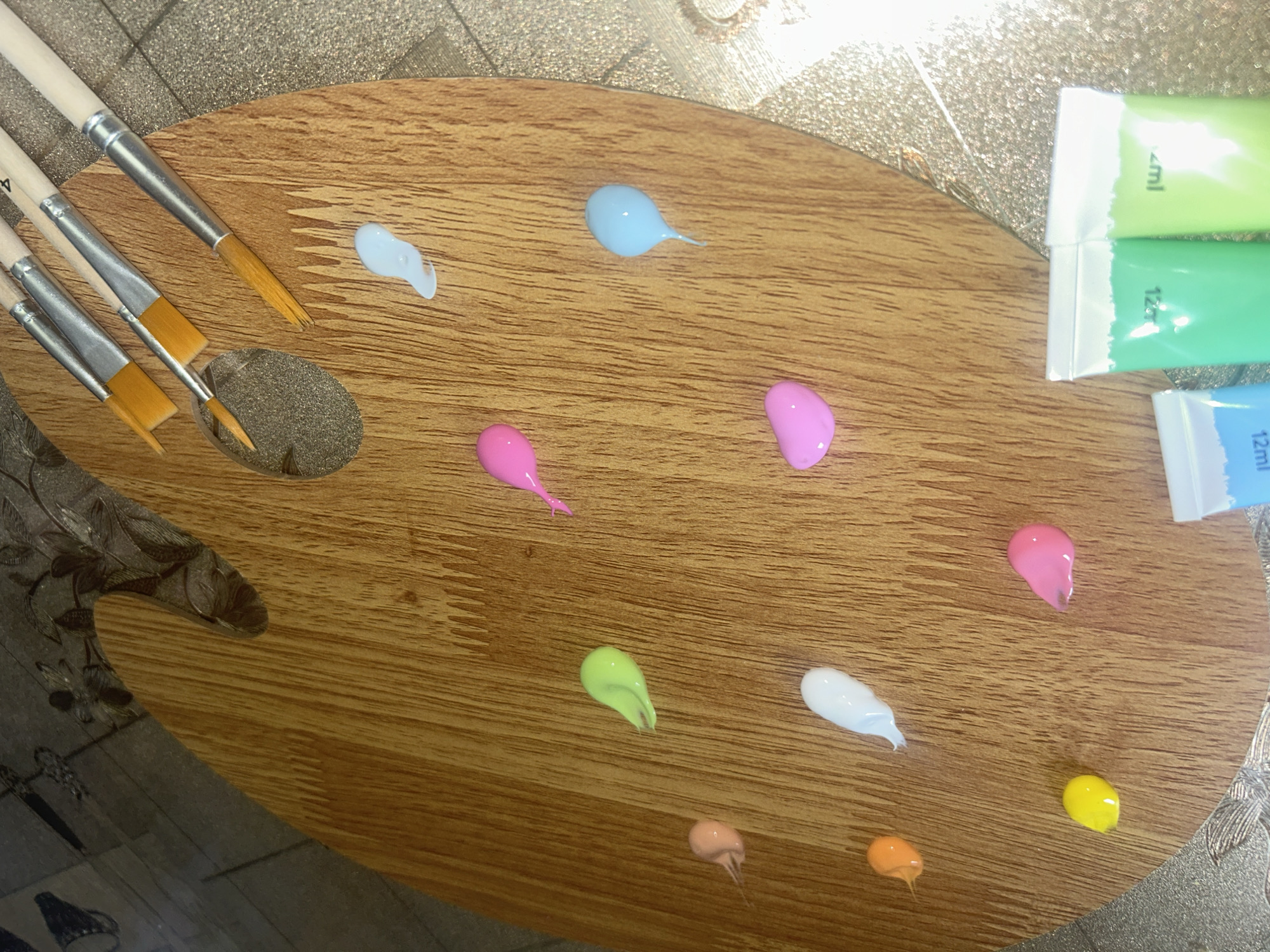 The image shows an artist's wooden palette with multiple small dollops of paint in various colors: light blue, blue, pink, light pink, lime green, white, peach, orange, and yellow. To the left of the palette are four paintbrushes. To the upper right of the palette are three partially visible paint tubes in green and blue. The palette is placed on a textured surface, possibly a table. The image provides no information about Dragovishtitsa, Bulgaria.