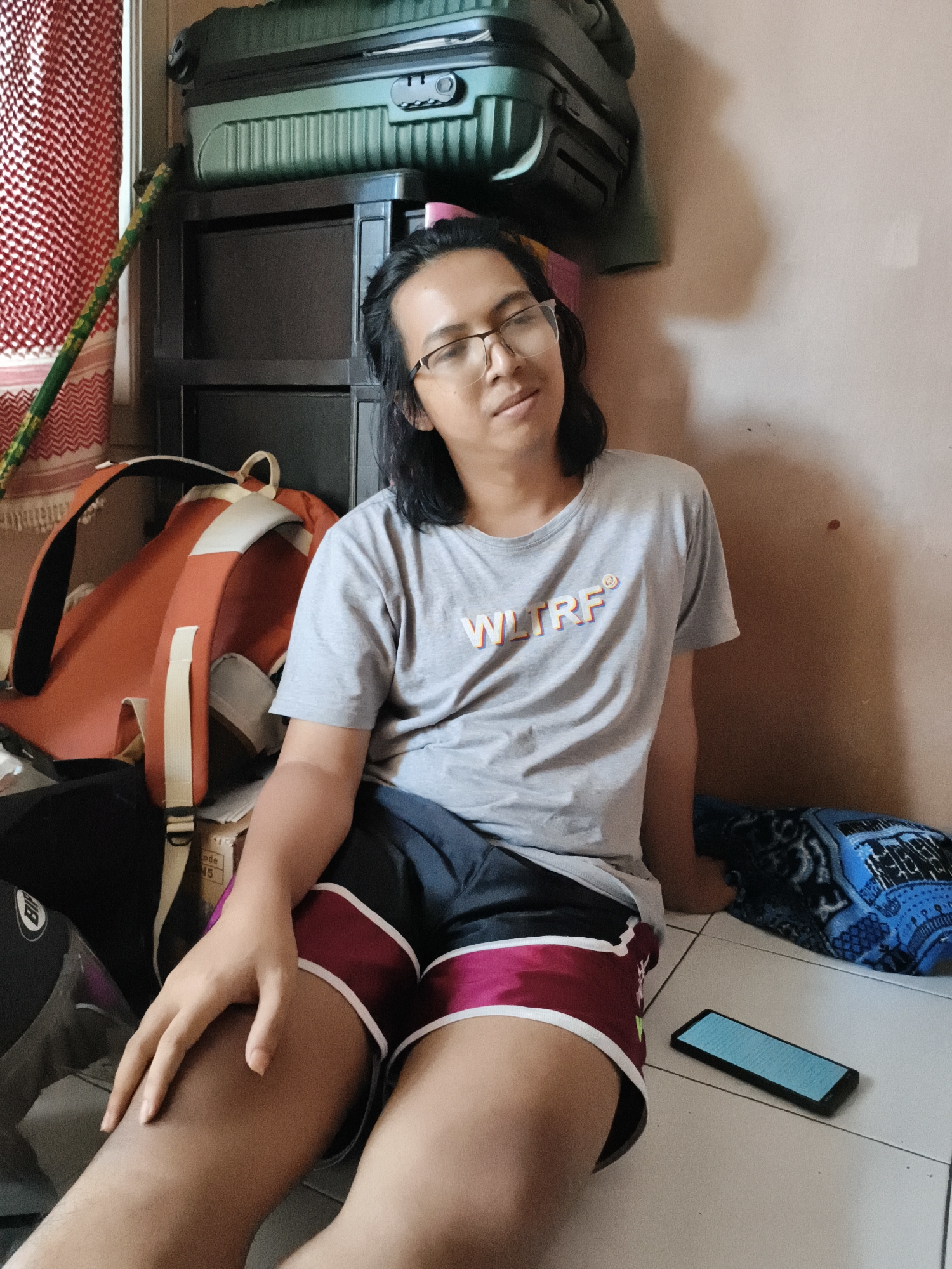 The image depicts a person sitting on the floor indoors, possibly in Cianjur, Indonesia, given the context. The individual has long dark hair, wears glasses, a grey t-shirt with the text "WLTRF" and dark shorts with pink and white trim. They are positioned near a collection of luggage, including a large green suitcase stacked on top of black drawers. An orange backpack is also visible. To the right of the person, a blue blanket and a smartphone lie on the floor. The background consists of a plain wall and a red and white patterned curtain on the left. The lighting suggests an indoor environment, possibly during the day. The person's posture and facial expression are relaxed, with their head tilted slightly upwards and their hand resting on their thigh.