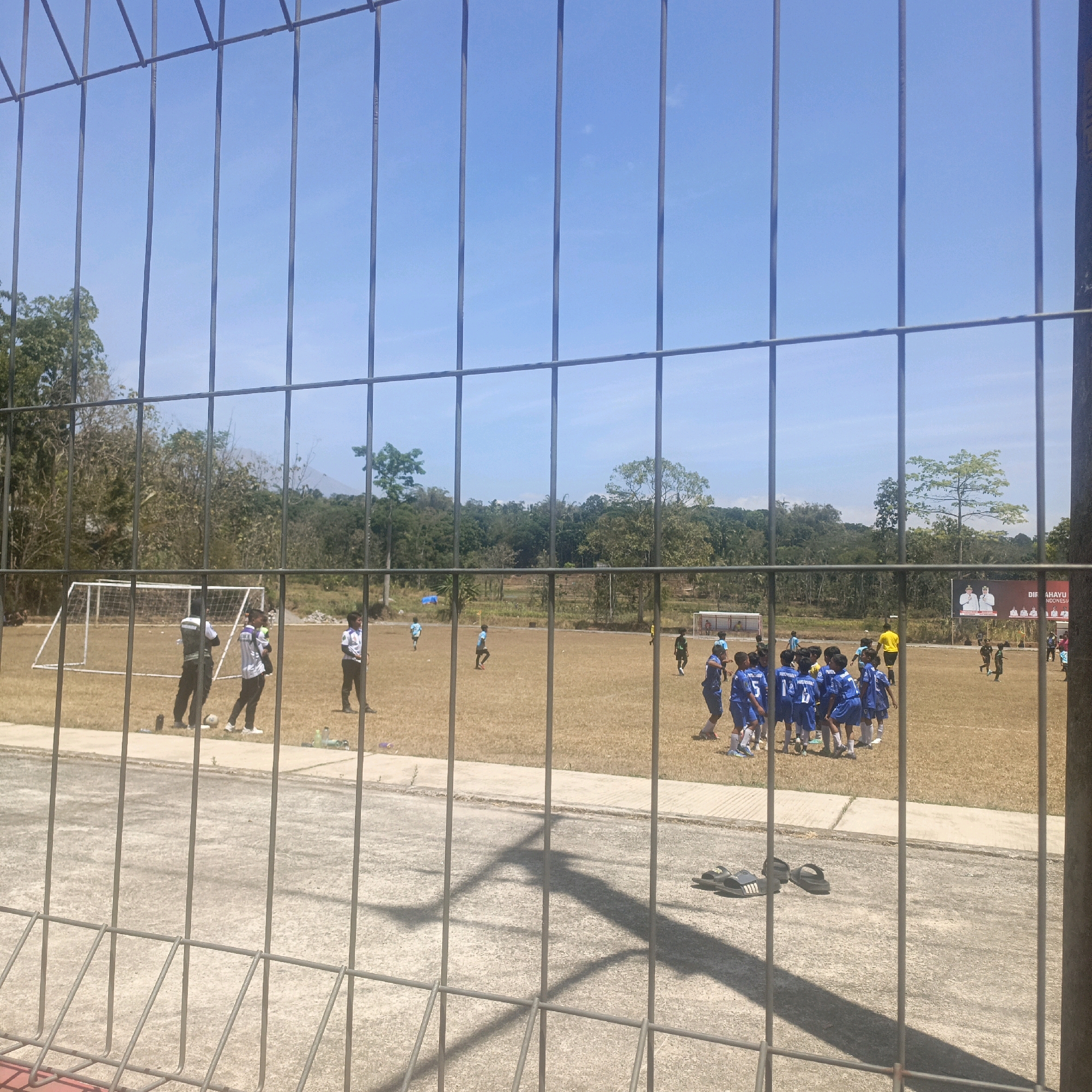 This image, captured in Krajan Lor, Indonesia, depicts a football field during daylight. A wire fence occupies the foreground, casting shadows on the concrete path beyond. On the field, several young boys in blue football uniforms are gathered, appearing to be engaged in a game or practice. A few adults, including one in a dark vest and another in a white shirt, are also present, possibly coaches or observers. A football goal is visible on the left side of the field, and trees and foliage form the backdrop. A large banner with images of people and text, likely promotional or informational, is displayed on the right side of the field. The sky is clear and blue.