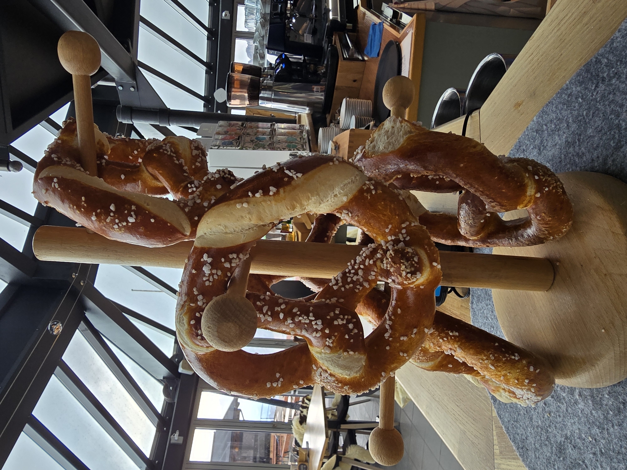 The image shows a wooden stand with several large, salted pretzels hanging from it. The stand is located in what appears to be a cafe or restaurant with a glass ceiling and a view of an outdoor area. In the background, there is a coffee machine, tables, and chairs. The pretzels are golden brown and sprinkled with coarse salt. The lighting is natural, coming from the glass ceiling. There are no people visible in the image. The overall scene suggests a cozy and inviting atmosphere. The location is identified as being in Garmisch-Partenkirchen, Germany.
