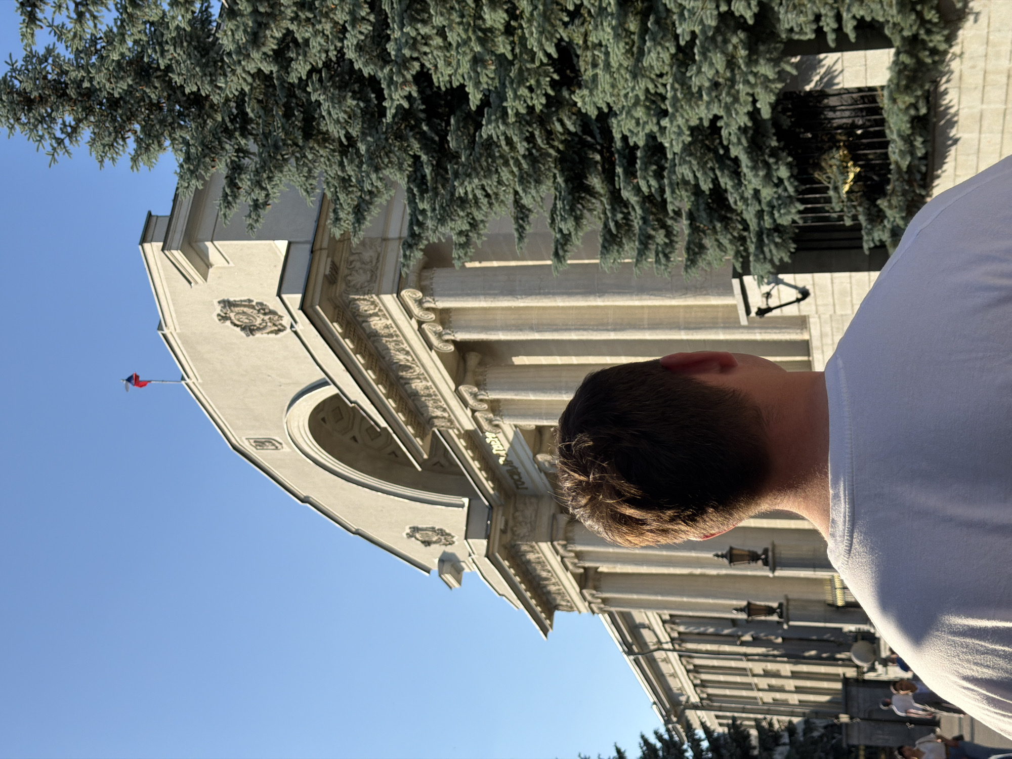 The image displays a rear-view perspective of a male subject wearing a white t-shirt, facing a large, classical-style building under a clear blue sky. The building, identified as the State Council of the Republic of Tatarstan, is situated in Kazan, Russia. It features light-colored stone, numerous prominent Corinthian columns, a large central archway, and intricate decorative reliefs. An inscription "ГОСУДАРСТВЕННЫЙ СОВЕТ" (State Council) is visible above the columns. A Russian flag (white, blue, red) flies from a flagpole on the building's roof. To the right, a large coniferous tree with blue-green needles partially obscures the building's facade. Additional evergreen trees are visible on the far left. Sunlight illuminates the scene, casting shadows. Several other individuals are visible walking in the background on the left side of the frame, near ornate street lamps.