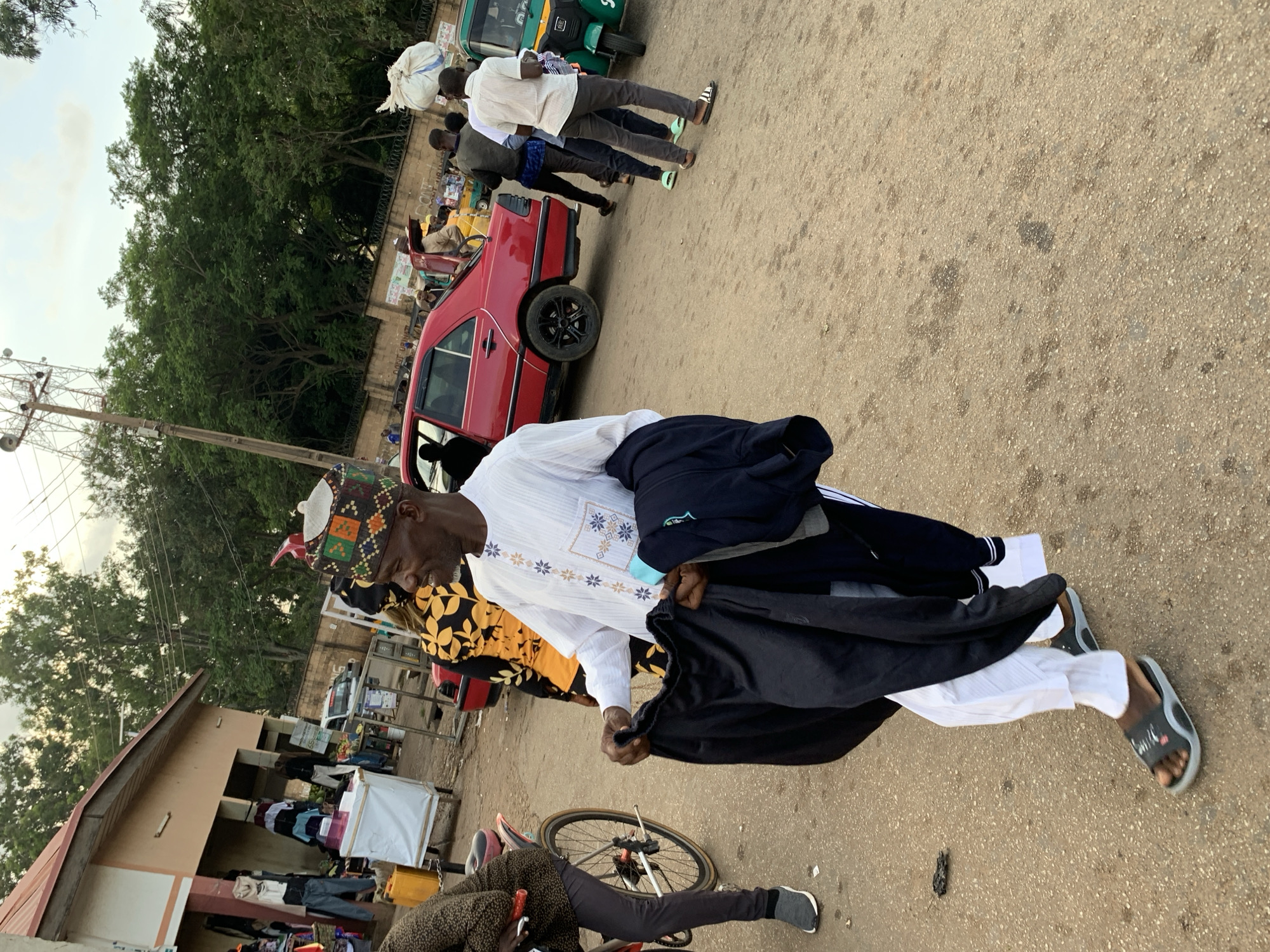The image shows a street scene in Jos, Nigeria, likely a market area. In the foreground, a man is walking and holding folded dark clothing. He is wearing a white garment with blue and gold embroidery and a patterned cap. He has a dark beard and is looking to his right. To his left, a person on a bicycle is partially visible. In the background, a red sedan is parked, and several other people are standing and walking. There are market stalls and trees in the background, with a communication tower visible in the distance. The ground is paved with asphalt, and there are some scattered debris. A green auto-rickshaw is also visible.