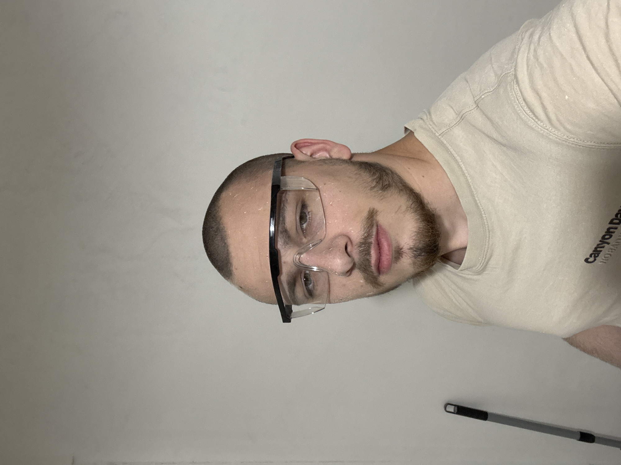 A close-up, tilted portrait captures a male subject indoors, likely in Stara Zagora, Bulgaria. The man, with short hair and a beard, is wearing clear safety glasses, which have some condensation or smudges on them. White speckles, consistent with paint splatters, are visible on his forehead and cheek, suggesting he is engaged in a painting or renovation project. He looks directly at the camera with a neutral expression.
He wears a beige crew-neck t-shirt. Partially visible text on his shirt includes "Canyon Dav..." and "NOBA", with some smaller, illegible text below it. In the bottom right corner of the frame, the handle of a paint roller pole is visible, further supporting the interpretation of a painting activity. The background consists of a plain, light-colored wall, possibly white or a very light neutral tone. The lighting appears even, suggesting an indoor setting without strong natural light cues for the time of day.