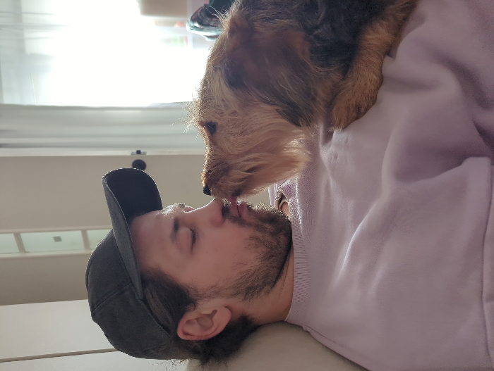 An adult male and a dog are depicted in an indoor setting, presumably a private residence in Moscow, Russia. The man, seen in profile from the chest up, occupies the left side of the frame, leaning back against a light-colored surface. He wears a dark grey baseball cap, reversed, and a light purple crew-neck sweatshirt. His eyes are closed, and his lips are puckered. A small, multi-toned brown and black shaggy-haired dog rests on the man's upper chest/shoulder, facing left. The dog's nose is making direct contact with the man's puckered lips. The background on the left consists of a plain, light-colored wall. To the right, a brightly illuminated window or doorway with light-colored curtains is visible, alongside a dark door handle on a white frame.