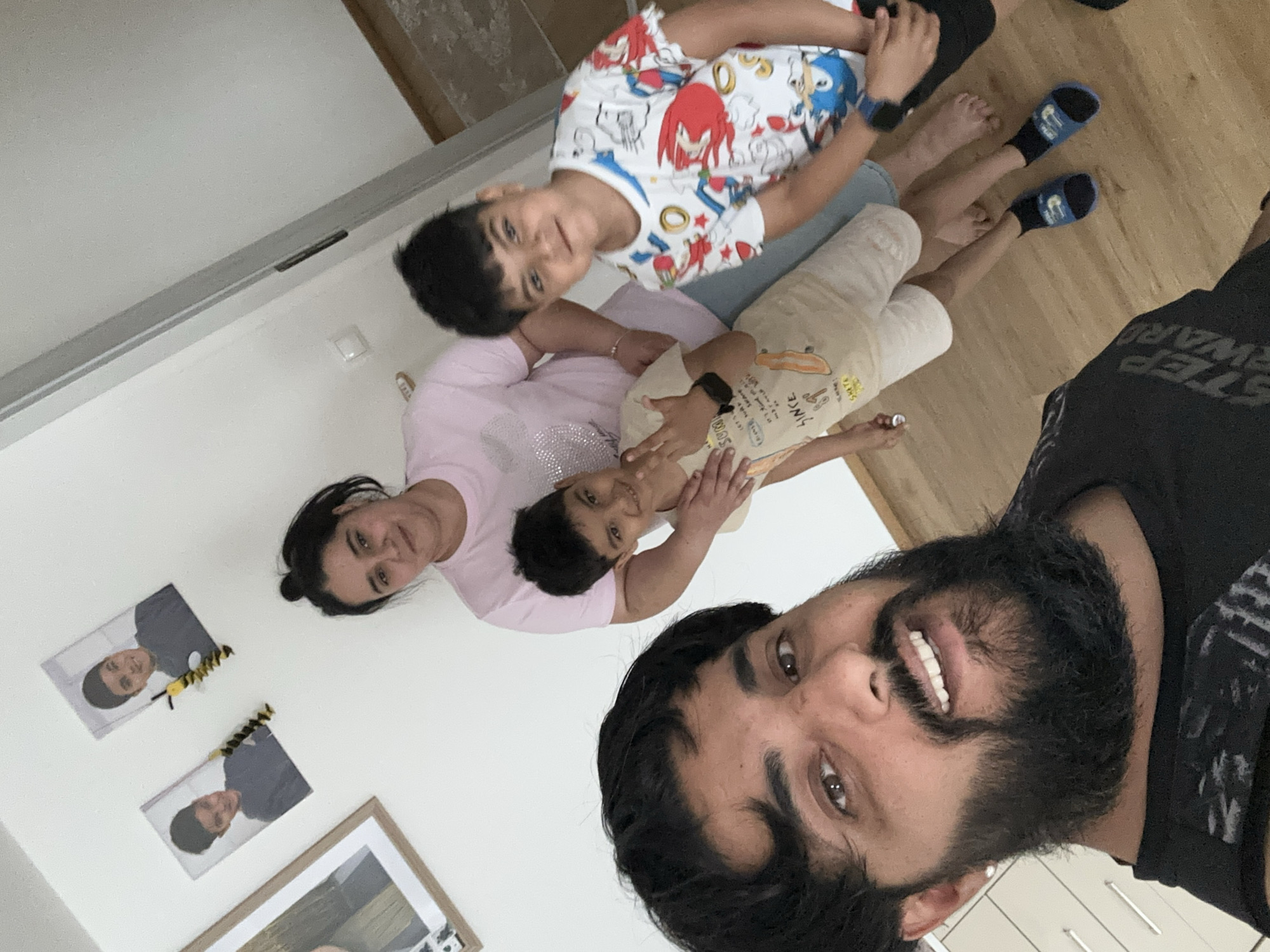 In a room with white walls and light-colored wooden flooring, a group of four individuals is captured in a selfie. The foreground is dominated by the close-up of a man with a thick, dark beard and brown eyes, smiling broadly. Behind him, a woman with dark hair pulled back in a bun wears a light pink t-shirt. Two young boys are also present. One boy, on the left, wears a light beige t-shirt and shorts, and is making a peace sign with his fingers. The other boy, on the right, is wearing a white t-shirt with a colorful cartoon print. The background shows framed pictures on the wall, including two portraits of a young boy and a decorative bee figure. The lighting suggests an indoor setting, possibly a home. The presence of framed family photos and the casual attire of the individuals indicate a personal and informal moment.
