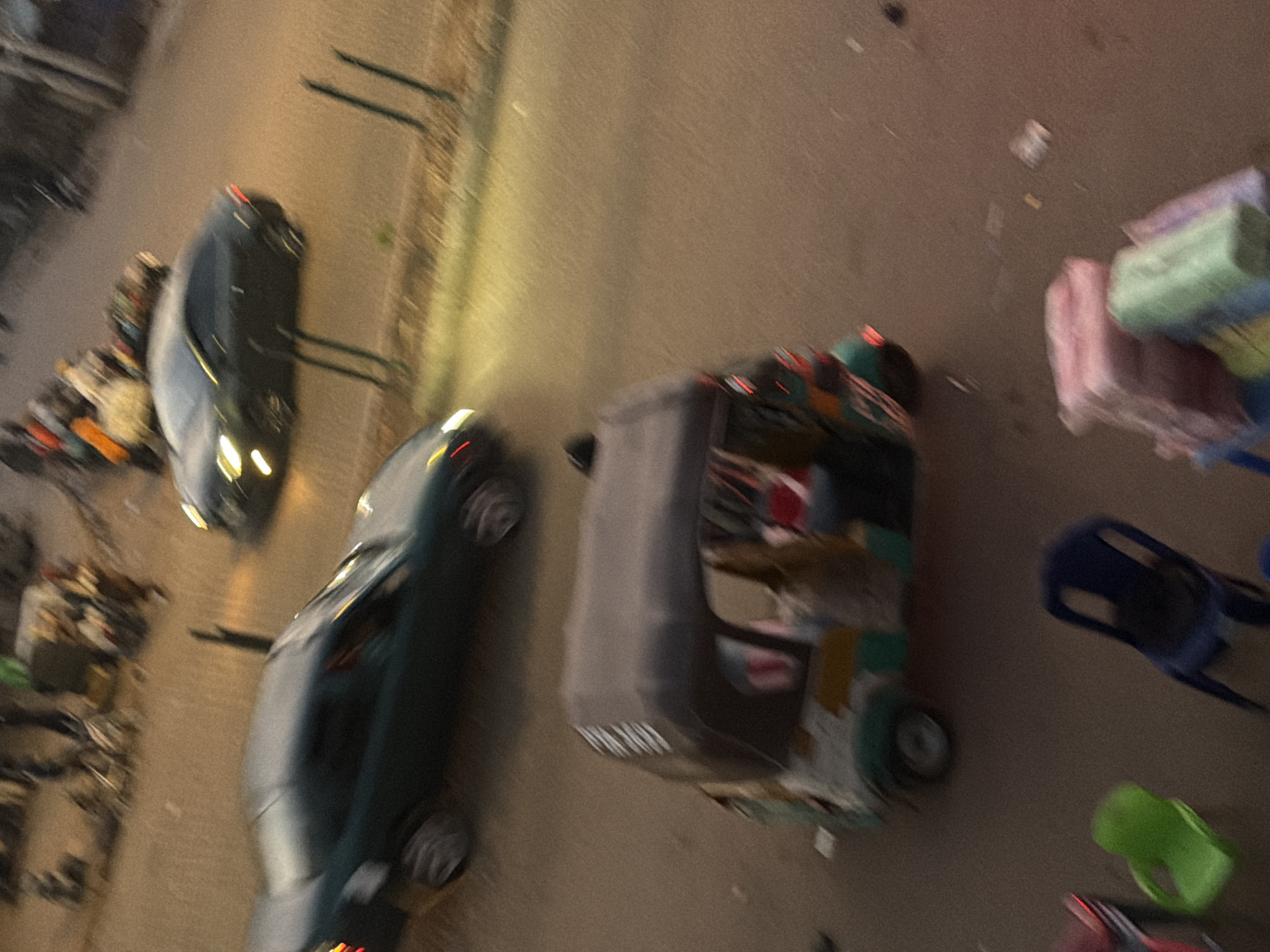 This is an aerial, blurry photograph taken at night in Bukuru, Nigeria. The scene appears to be a street with several vehicles and miscellaneous objects. In the foreground and to the left, a dark-colored car is visible, with its headlights on. To its right, a tuk-tuk or auto-rickshaw is overturned, its wheels pointing upwards. Further to the right, several stacks of goods wrapped in plastic are piled up, one appearing to be green and others pink. In the background, another dark-colored vehicle is partially visible, along with what look like streetlights. Scattered on the ground are various small objects, possibly debris. The overall impression is of a busy street scene at night, with the overturned vehicle suggesting a possible accident or disruption. The low light conditions and motion blur contribute to a sense of urgency or chaos.