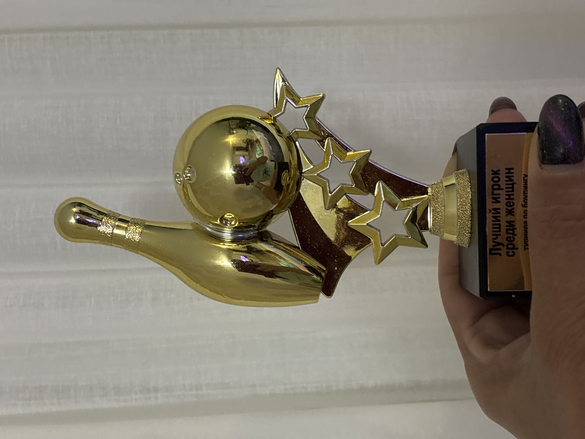 A person's hand, with dark, glittery nail polish, holds a golden bowling trophy. The trophy features a bowling pin and ball, accented by star shapes and a black base with an inscription in Russian that reads "Лучший игрок среди женщин турнира по боулингу" (Best female player in the bowling tournament). The background is a light-colored, textured curtain. The reflection in the bowling ball shows a person holding a phone, likely taking the picture, and a room interior.