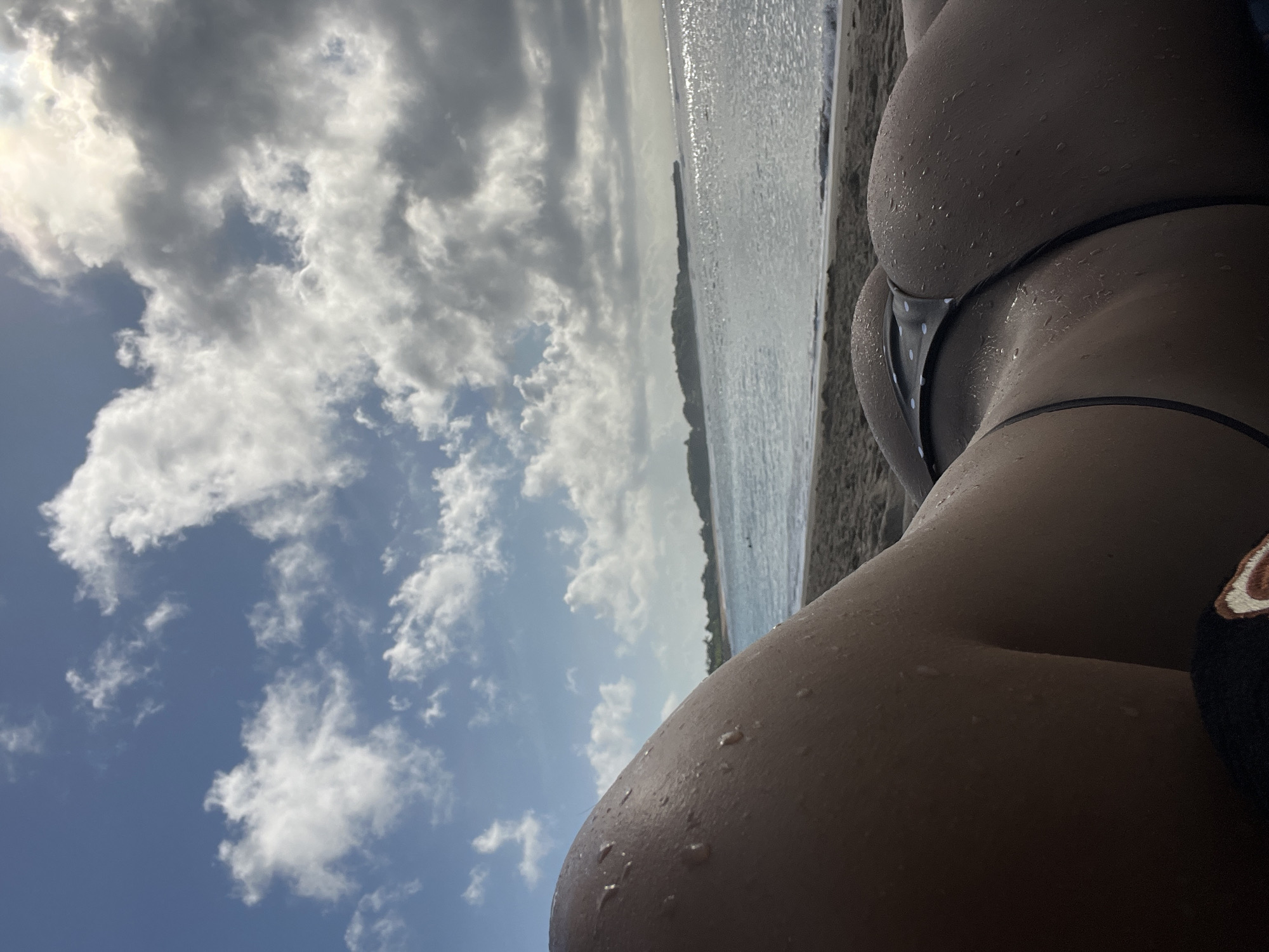 A photograph taken from a low angle, slightly tilted, shows a person's buttocks in a polka-dotted bikini bottom on a beach. Water droplets cling to the skin. To the left, the background features a blue sky filled with fluffy white clouds. In the distance, a body of water, likely the ocean, stretches towards the horizon, where a line of vegetation is visible, possibly indicating the coastline. The photograph was likely taken in Kuta Selatan, Indonesia.