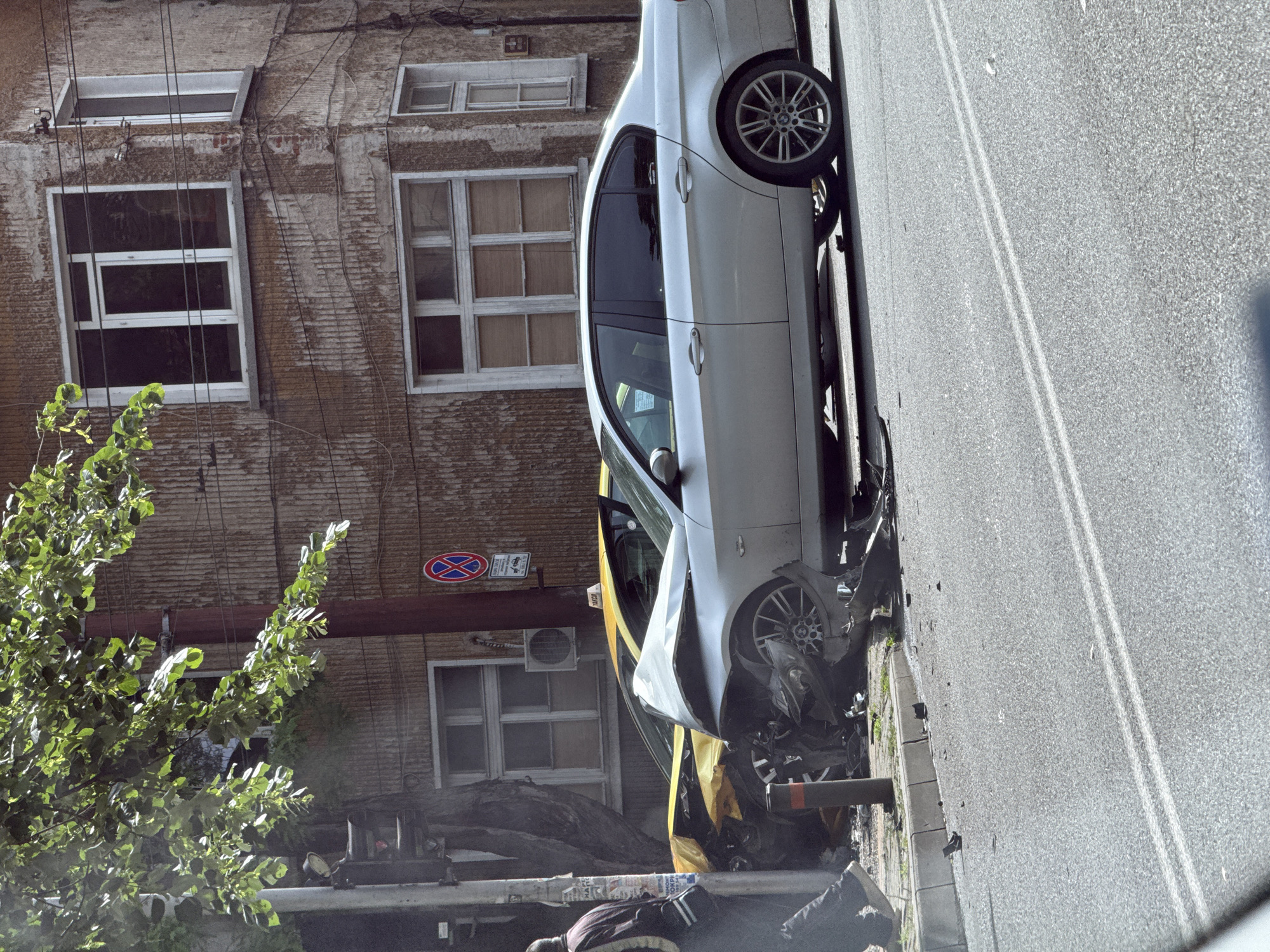 The image depicts the aftermath of a car accident in Stara Zagora, Bulgaria. A white car, severely damaged at the front, rests on the sidewalk against a metal bollard. A yellow car is partially visible beneath the white car, also indicating collision involvement. A man lies on the ground near the wrecked vehicles, possibly injured. The accident occurred on a street with double white lines. A weathered building with windows is visible in the background. A no-parking sign is attached to the building.
