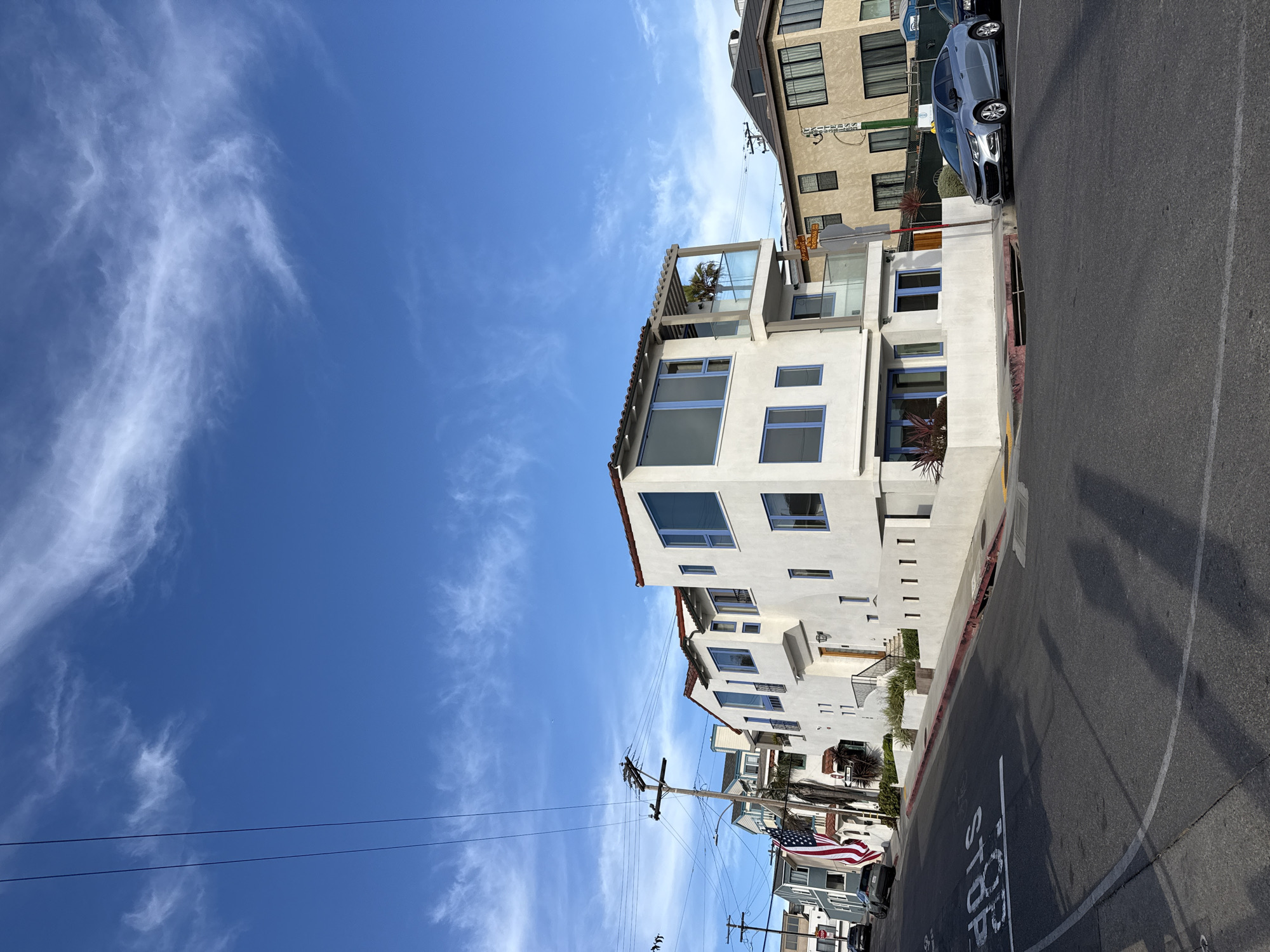 This is a medium-distance, eye-level, outdoor, full shot of a residential street corner in Hermosa Beach, California, USA. The sky is a clear, bright blue with wispy white clouds. A power pole with overhead wires is visible on the left side of the frame. The street is paved, with a white stop line painted on it in the foreground.
The primary subject is a modern, white, three-story house with blue-trimmed windows and a flat roof. An American flag is displayed on a pole to the left of the house. Adjacent to the white house is a tan, three-story building. A silver sedan is parked on the street in front of the tan building. Another dark-colored vehicle is parked further down the street on the left. Street signs are visible on a pole near the intersection. The overall scene depicts a sunny day on a residential street with distinct architecture.