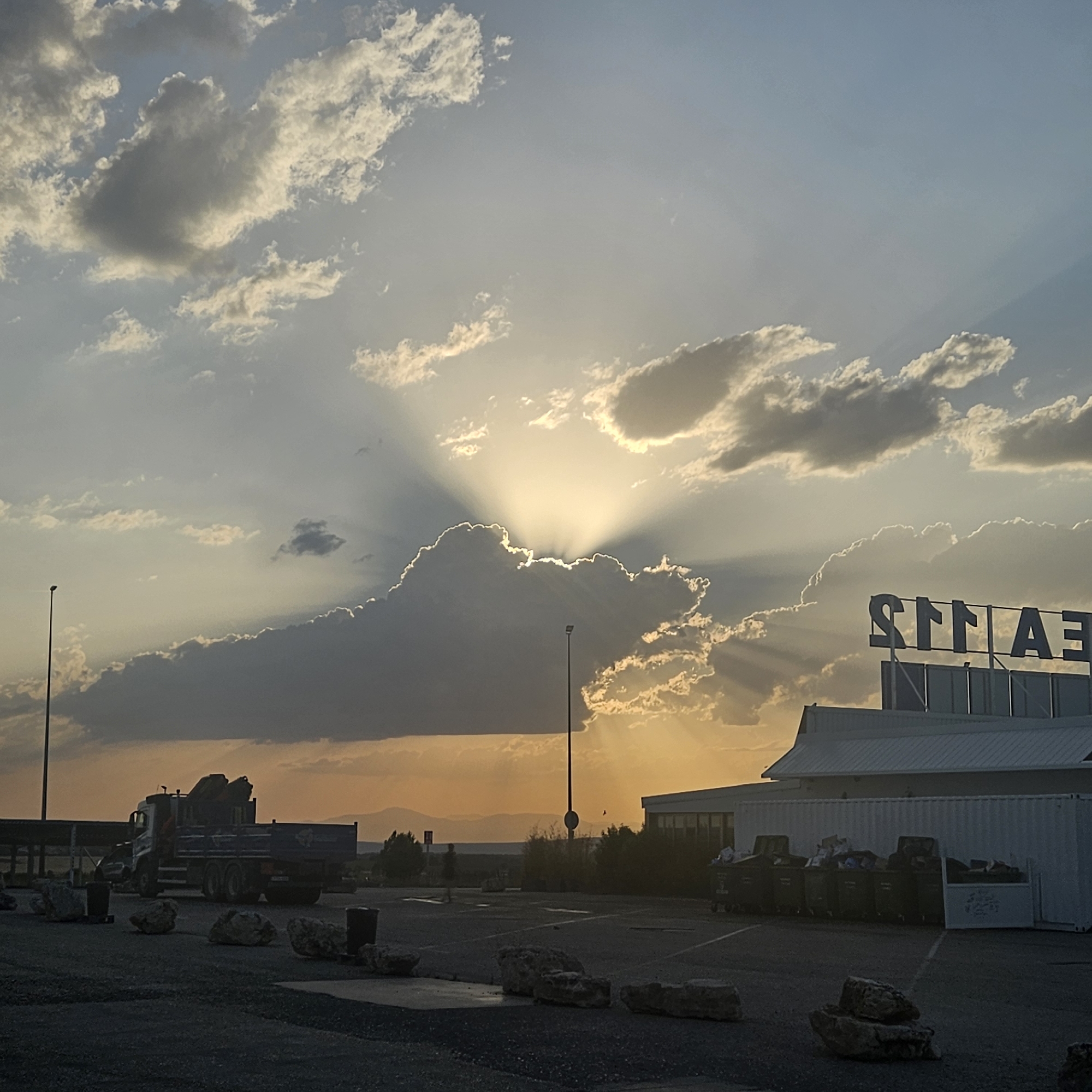 The image depicts a sunset scene in Algora, Spain. The sky is filled with dramatic clouds, with sunlight streaming through a large, dark cloud formation in the center, creating visible sunbeams. In the foreground, a paved area, possibly a parking lot, is visible, with several large rocks and some dark bins scattered across it. A large truck with a crane attachment is parked on the left side of the frame. In the background, a modern building with a prominent sign featuring large, stylized letters that appear to read "STIAS" or a similar configuration is visible on the right. The overall atmosphere is characterized by the warm, golden light of the setting sun.