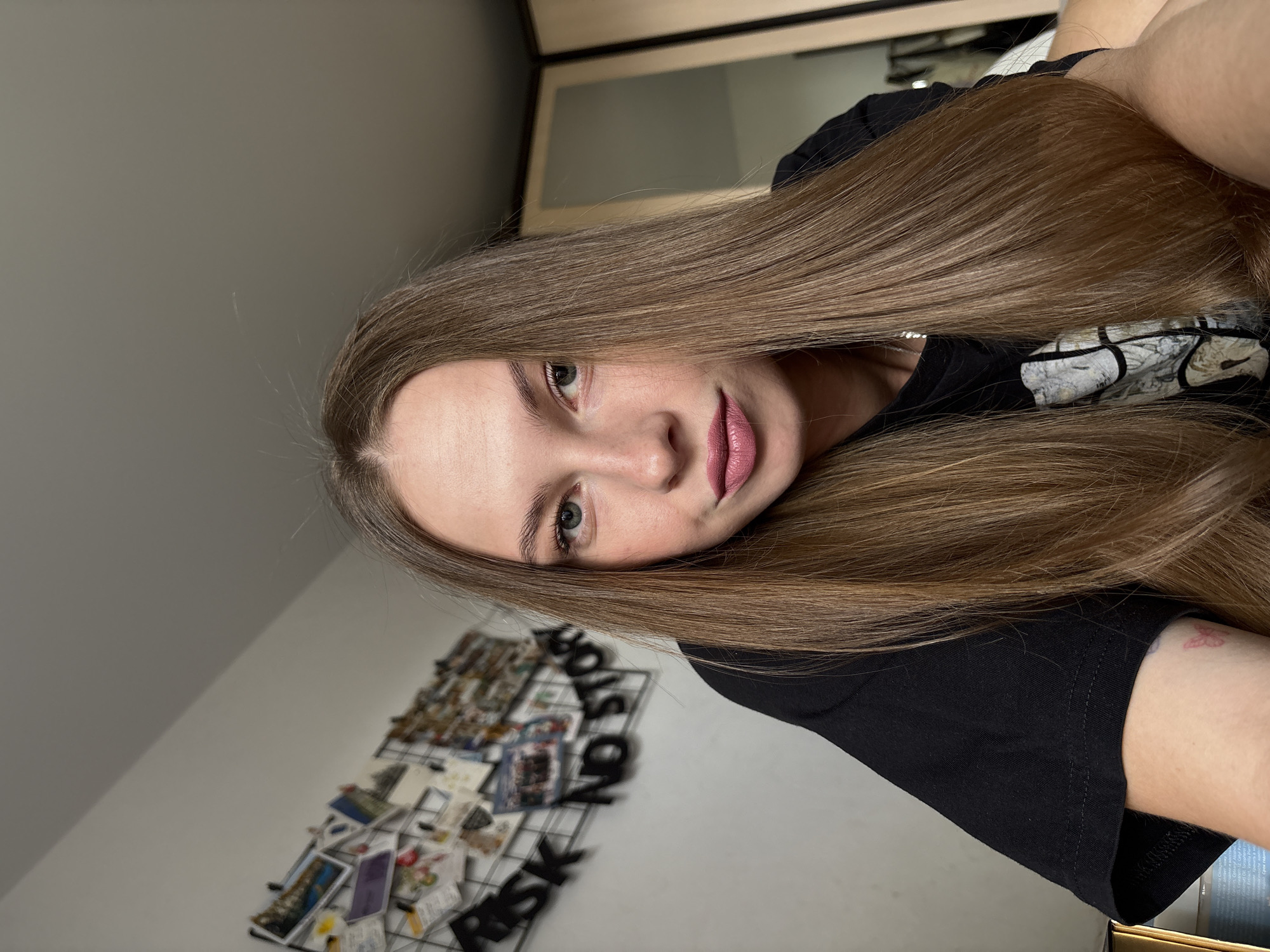 A young woman with long, straight, brown hair is shown in a close-up, eye-level shot, looking directly at the camera. She is wearing a black t-shirt and her lips are lightly colored. The background is a plain white wall with a bulletin board covered in photos and papers. Black letters spelling out "RISK NO STORY" are attached to the bulletin board. The environment appears to be indoors, possibly a bedroom, with natural light coming from the right. The overall impression is informal and personal. The image does not contain any specific geographical markers that would indicate a location in Perm, Russia.