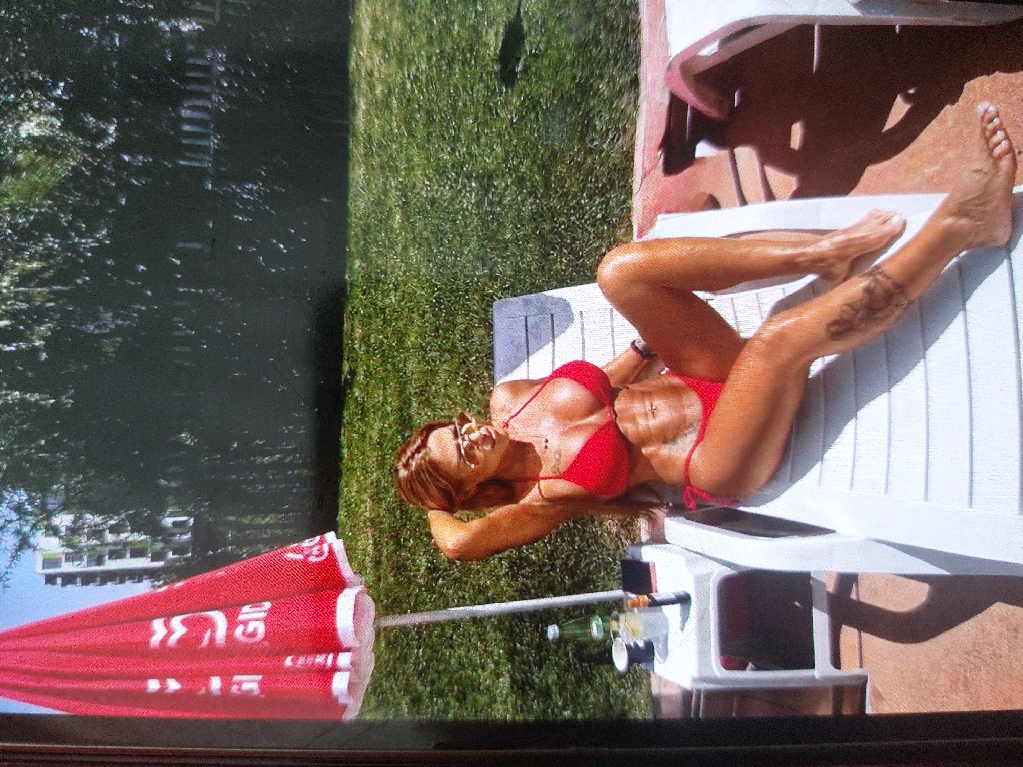A woman is lying on a white lounge chair outdoors. She is wearing a red bikini and sunglasses. She is on a grassy area next to a building. A red umbrella is nearby. The location is Tsarevo, Bulgaria.