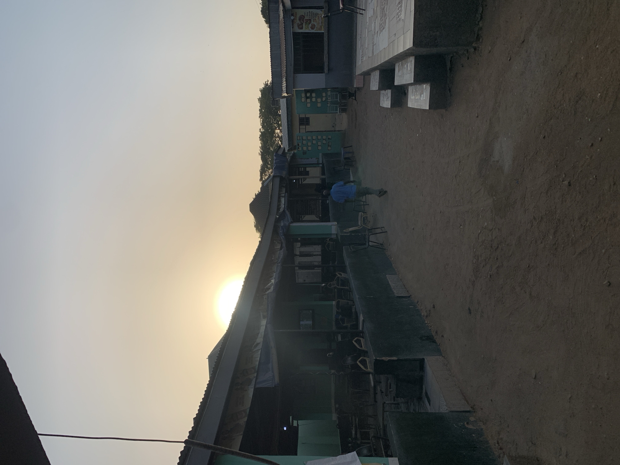 The image depicts an outdoor scene at New Stadium, Nigeria, at what appears to be a restaurant or roadside eatery. The sun is visible in the sky, indicating either dawn or dusk. The eatery is a simple structure with a corrugated iron roof and green-painted walls. A few individuals are present, seemingly patrons, seated at tables and chairs. The ground is mostly bare dirt. A second building or structure stands in the background, and there is a dirt pathway leading from it. The environment seems typical of a developing urban or semi-urban setting.