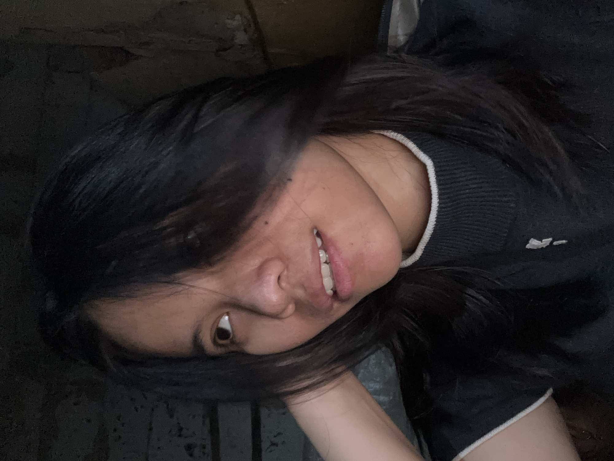 A close-up, waist-up, medium-shot, indoor photograph depicts a young woman with dark, shoulder-length hair, partially obscuring her face as she looks to her left. She is wearing a dark blue sweater with a white trim around the neckline. Her left arm is raised and resting behind her head. The background is dark and appears to be an interior wall with some texture, possibly brickwork. The lighting is dim, creating a dramatic effect. The image was taken in East Jakarta, Indonesia.