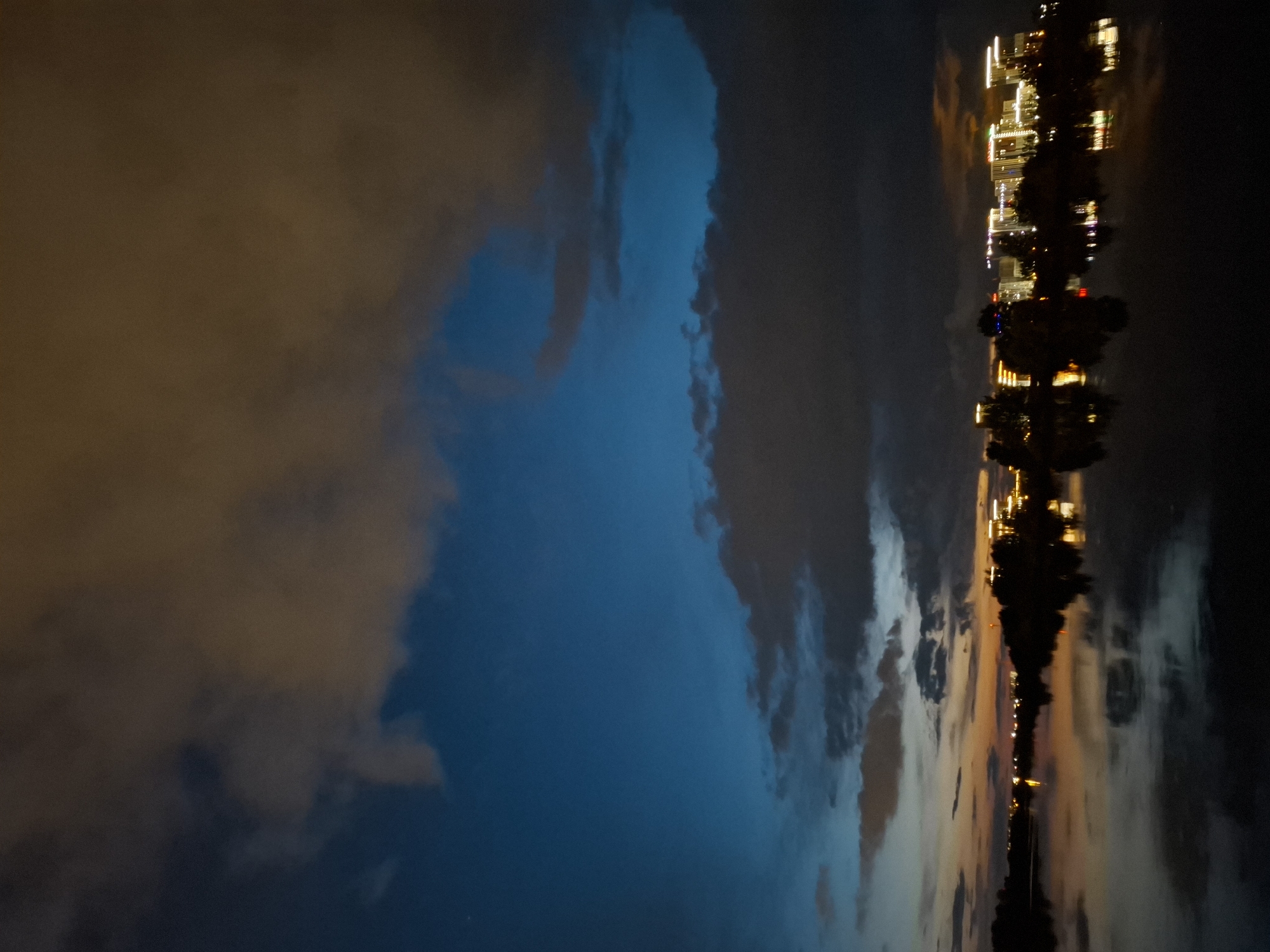 The image depicts a twilight scene over a body of water, likely a river or lake, with a city skyline in the distance. The sky is a mix of deep blue and darker clouds, with some lighter, warm-toned clouds reflecting the last vestiges of daylight. The water's surface is calm and reflects the lights of the city and the sky.
The city skyline consists of several modern buildings, illuminated with warm-toned lights, suggesting it is evening or night. These buildings are silhouetted against the sky, with some trees or foliage visible in front of them. The reflection of the lights and structures is clearly visible on the water.
The overall impression is one of urban tranquility at dusk. Without specific landmarks visible, pinpointing the exact location within Tyumen is not possible. However, such a scene is characteristic of waterfront areas within or adjacent to developed urban centers. The atmosphere is peaceful, with the gentle illumination of the city contrasting with the deepening blues of the sky. There are no people or active events visible in the image.
