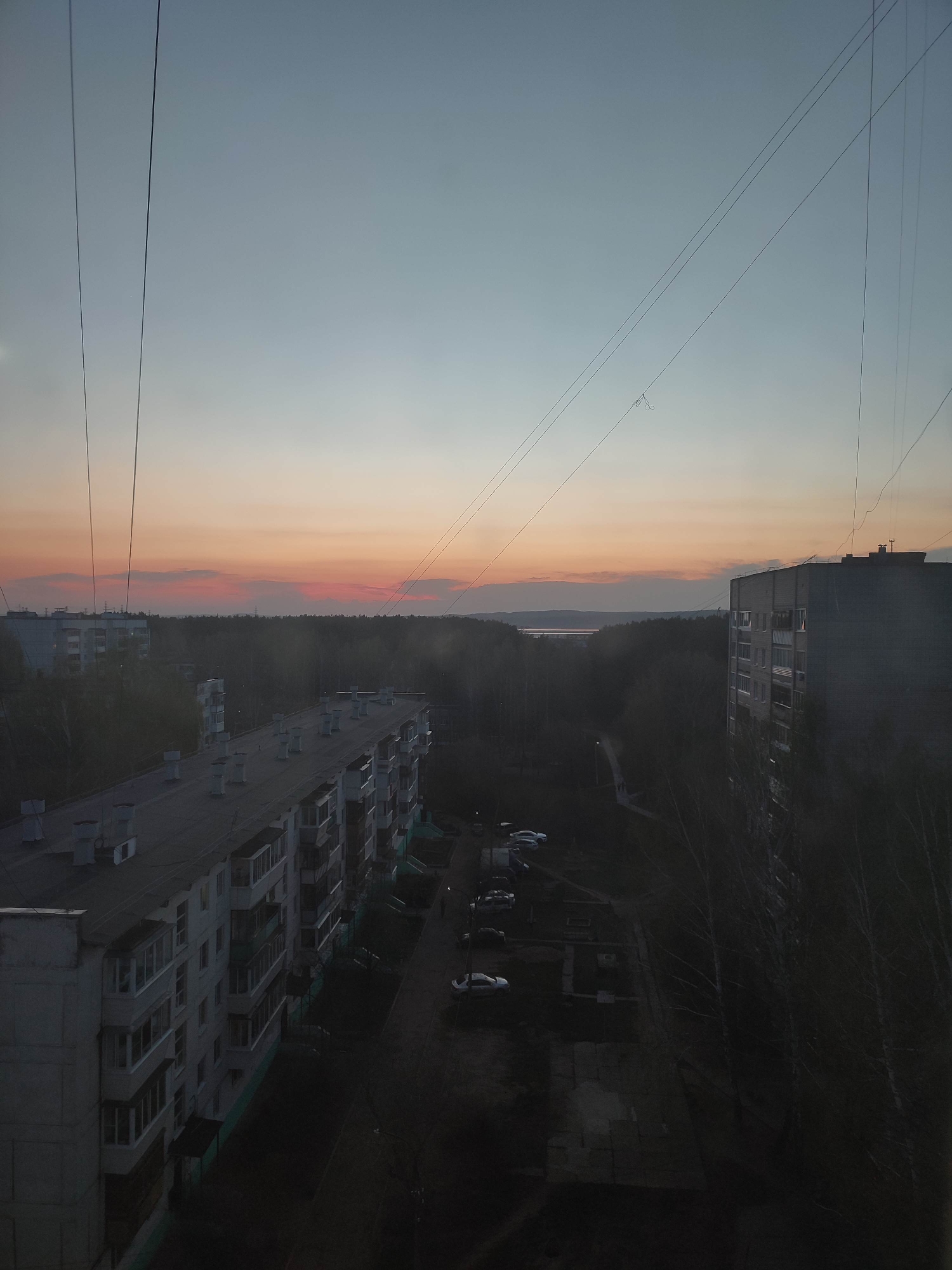 The image depicts an urban scene at dusk. Two multi-story apartment buildings, typical of Soviet-era architecture, dominate the foreground. The buildings are constructed of concrete, with multiple balconies and chimneys. A small parking lot with several cars is visible between the buildings. Beyond the buildings lies a dense forest, and in the distance, a body of water reflects the fading light. The sky is a gradient of blue and orange, indicating either sunrise or sunset. Power lines are strung across the sky, adding to the urban landscape. The overall mood is subdued and slightly gloomy.