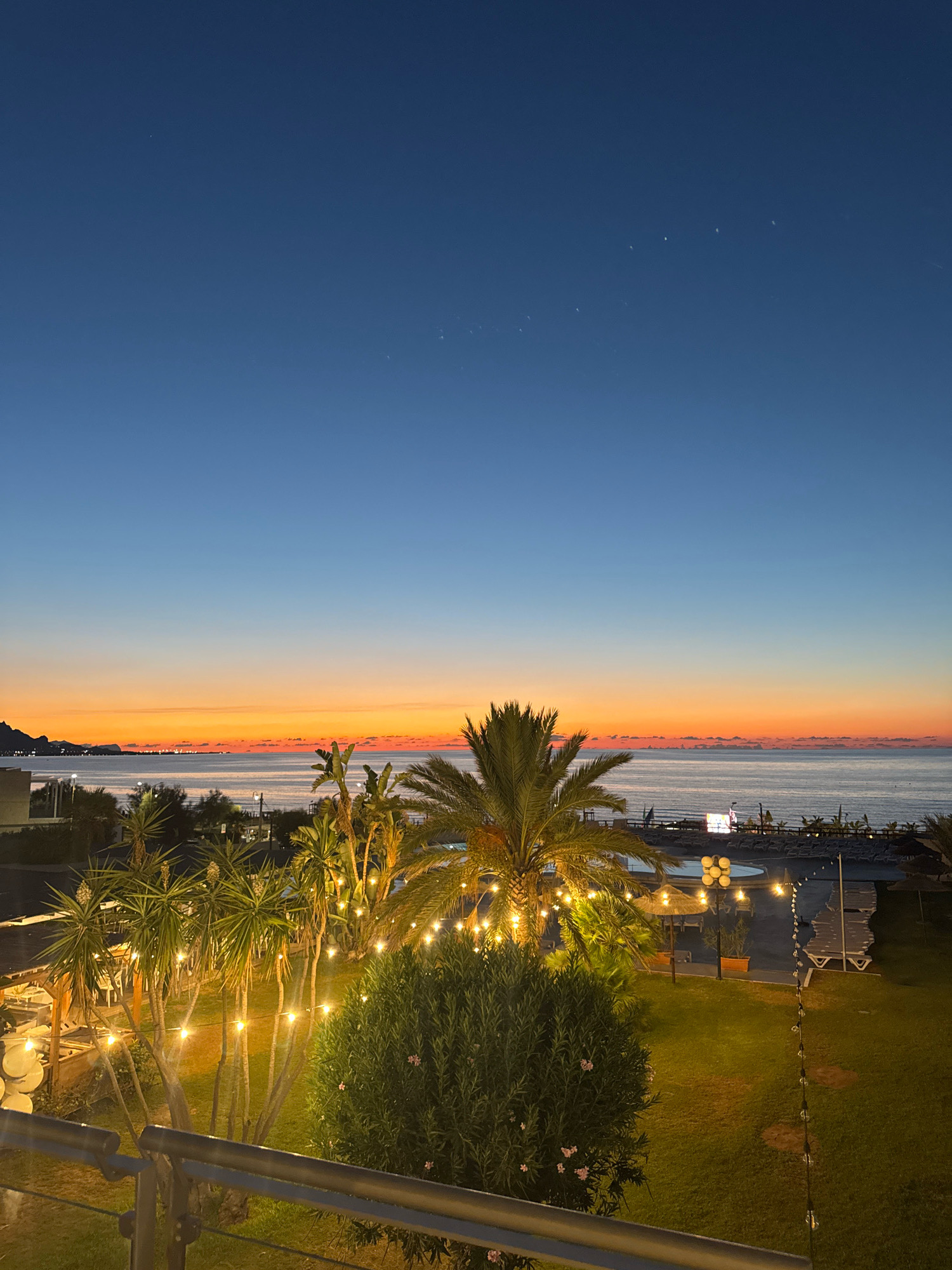 This is a vertical, medium-shot, outdoor photograph taken at dusk, likely from an elevated position such as a balcony, looking out over a coastal resort area in Isola delle Femmine, Italy. The sky transitions from a deep blue at the top to a gradient of orange and yellow near the horizon, indicating the last moments of sunset. The sea is visible on the horizon, with a faint line of lights on what appears to be a distant coastline and mountains.
In the foreground, there is a railing made of metal and wood. Beyond the railing is a well-manicured lawn. Several palm trees and other lush green foliage are scattered throughout the scene, many of them adorned with strings of warm, glowing fairy lights, creating a festive atmosphere. Some of these lights are also strung along a pathway or fence in the middle ground.
In the middle ground and background, a resort area is visible. This includes lounge chairs and umbrellas set up on what appears to be a beach or poolside area. A circular swimming pool is also present. The overall scene evokes a sense of tranquility and leisure, with the illuminated lights adding a magical touch to the twilight setting. There are no people clearly visible in the frame. The image captures the transition from day to night in a picturesque coastal location.