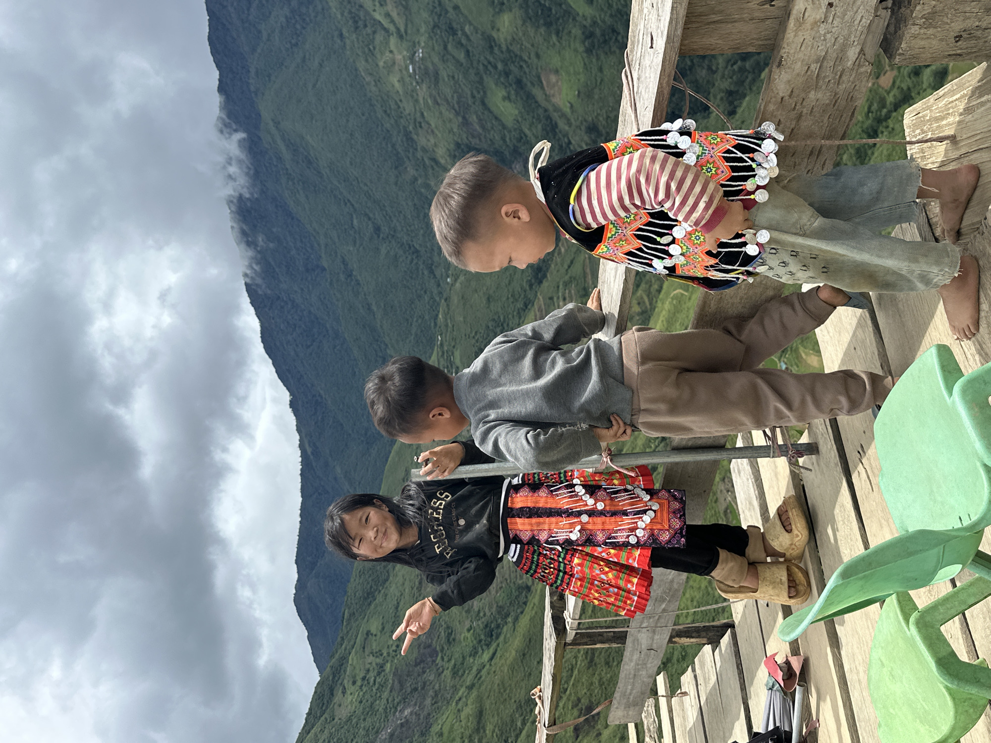 Three children stand on a wooden platform overlooking a lush, mountainous landscape in Bắc Yên, Vietnam. A young girl, center-left, smiles at the camera, holding up two fingers in a peace sign. She wears a black long-sleeved shirt, a brightly colored, patterned skirt adorned with decorative elements, and tan platform sandals. To her right, a young boy in a grey long-sleeved shirt and tan pants stands with his hands on his hips, facing away from the camera. On the right, another young boy, facing left in profile, wears a red and white striped long-sleeved shirt and a black vest with colorful embroidery and silver embellishments. He is barefoot and wears light-wash jeans. In the foreground, two bright green plastic chairs are visible. The background features rolling green mountains shrouded in a slightly overcast, cloudy sky. The wooden platform appears rustic, constructed from planks and supported by vertical posts.