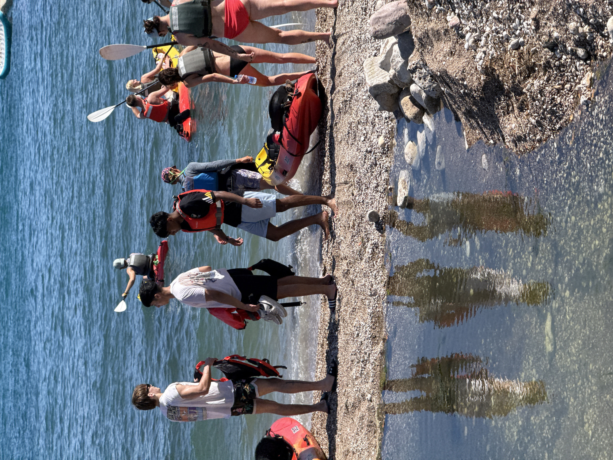A group of people are gathered on a pebble beach with kayaks near the water's edge. Two young men in the foreground are holding kayaks and bags. One man wears a white t-shirt and patterned shorts, while the other wears a white t-shirt and dark shorts. Several other individuals are visible in the background, some in kayaks on the water and others on the beach. The water is blue and appears to be calm. The scene is set during daylight hours, with clear sunny weather. The location is identified as Budva, Montenegro.