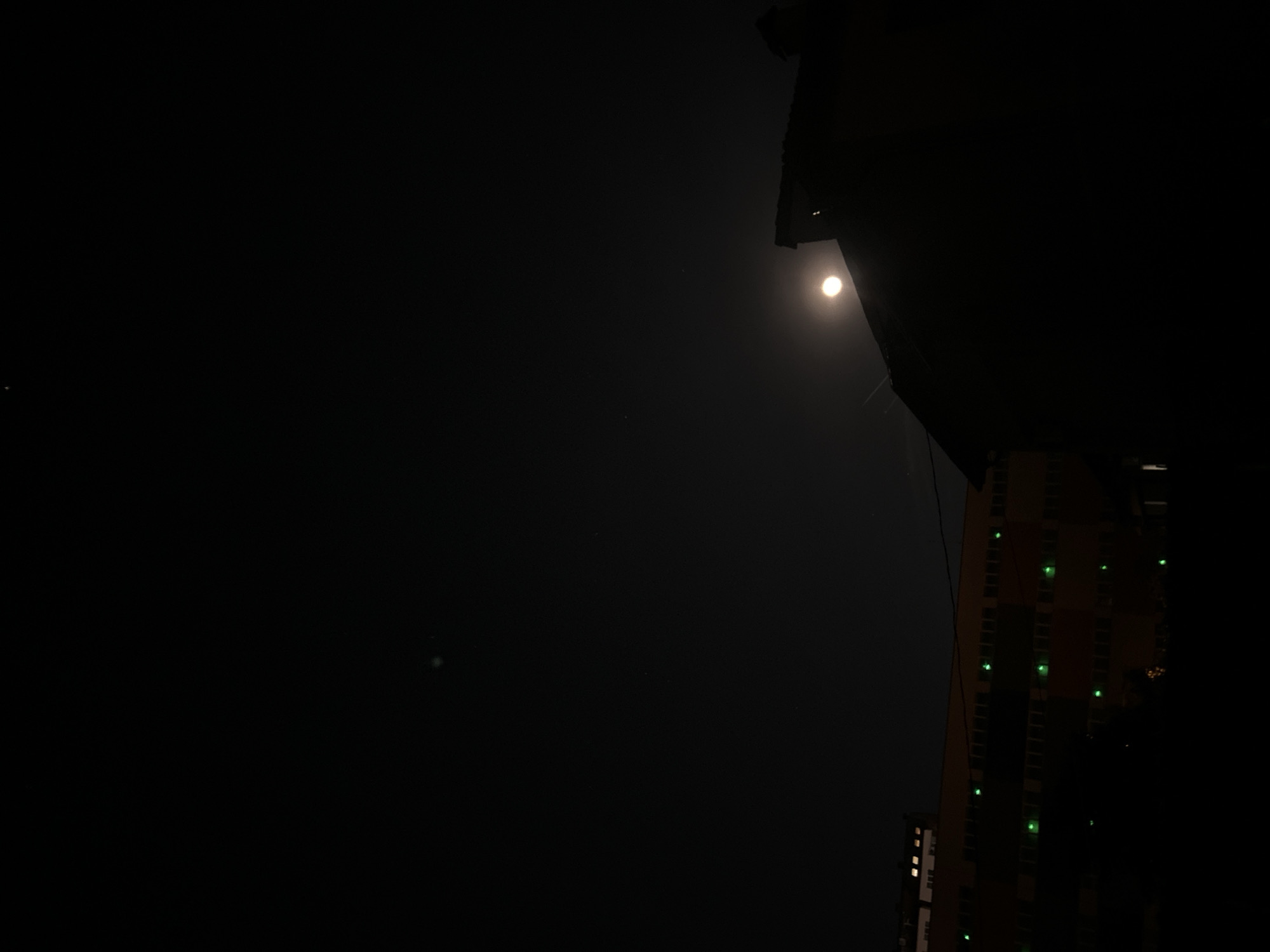 A dark, low-angle shot reveals a night scene in what appears to be Sofia, Bulgaria. A bright moon shines in the upper right, partially obscured by a dark structure, possibly the roofline of a building. Below and slightly to the right of the moon, a tall building is barely visible, with several floors lit by green lights. The rest of the image is mostly dark, indicating a night environment with limited ambient light.