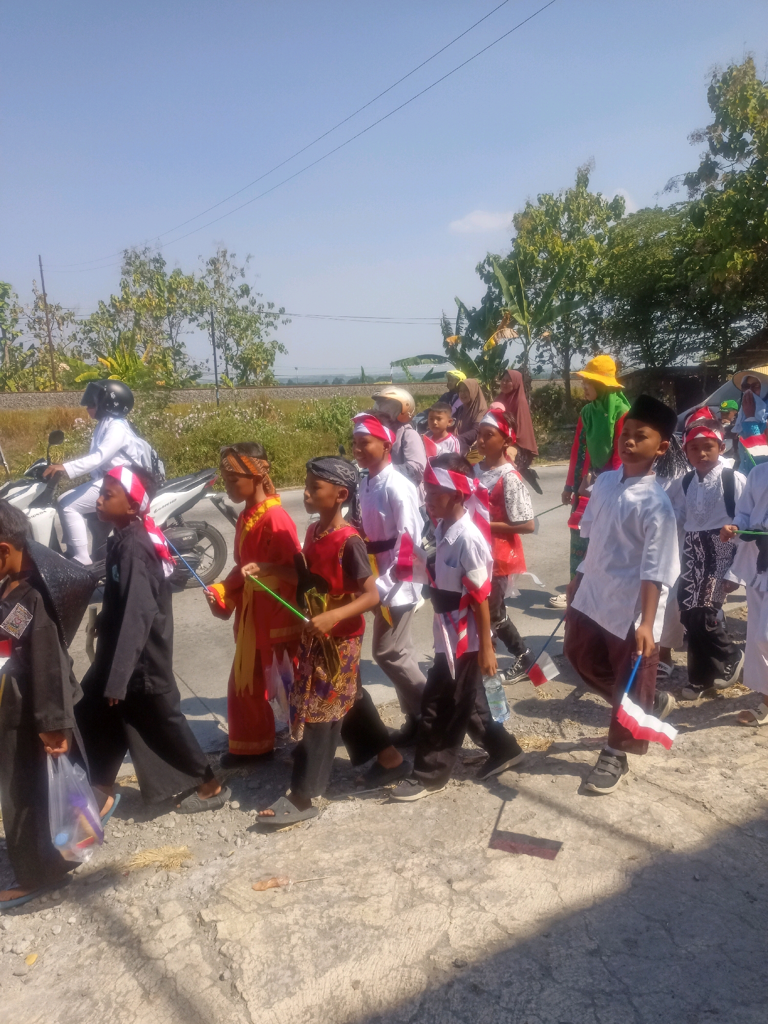 In the city of Sumberlawang, Indonesia, a procession of children walks along a paved road under a clear blue sky. The children, dressed in traditional Indonesian attire and various costumes, carry small Indonesian flags. Some children wear headbands with red and white stripes, symbolizing the national flag. One child in the foreground is dressed in a white shirt and brown sarong, holding an Indonesian flag. Another child to his left wears a red and yellow outfit with a yellow sash. Behind them, a person on a white motorcycle wearing a white outfit and a black helmet rides along the side of the road. Trees and foliage line the background, suggesting a rural or semi-rural setting. The ground is a mixture of asphalt and gravel. The overall scene depicts a cultural event or celebration.