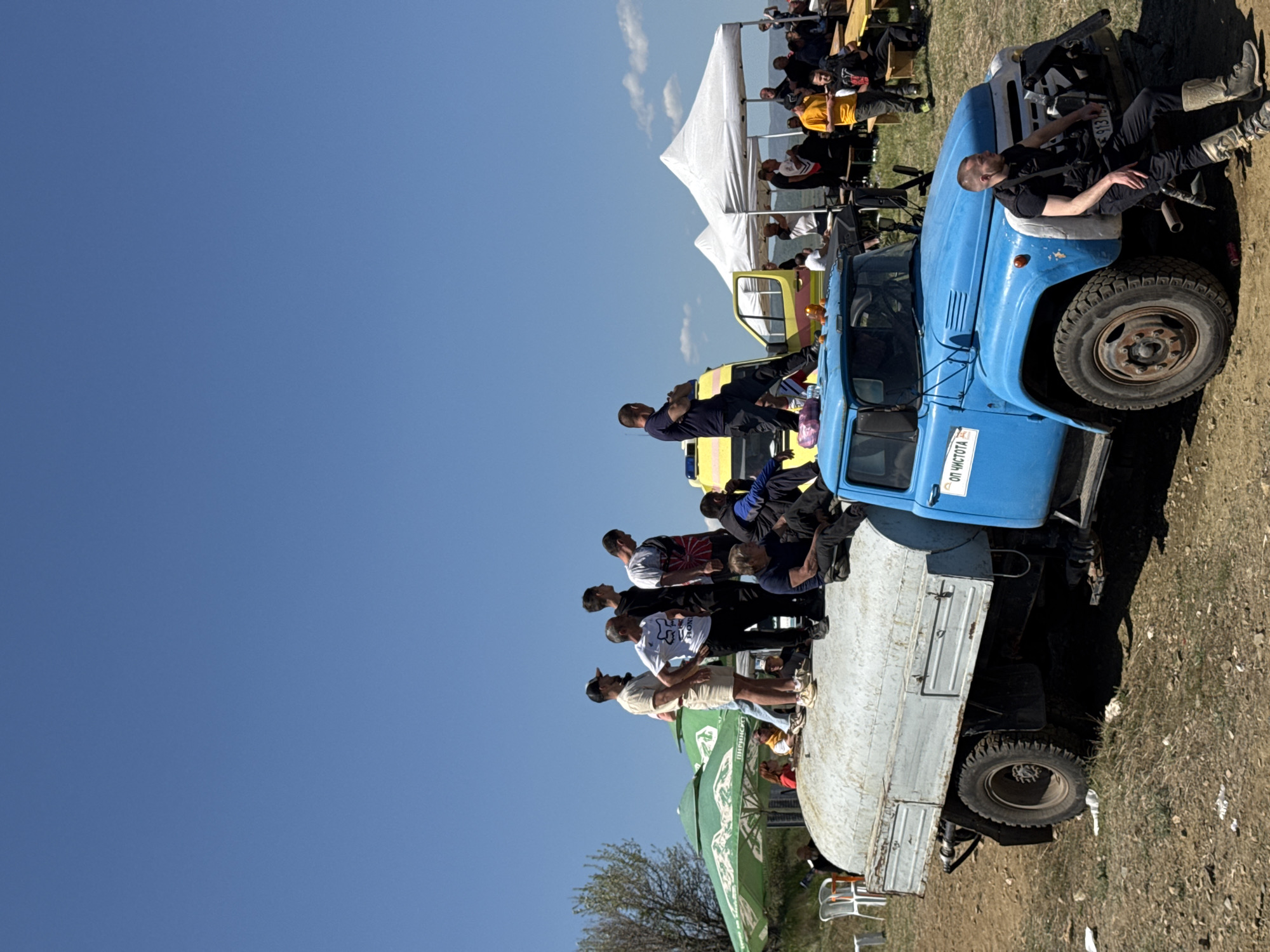 The image shows an outdoor event taking place in what appears to be a rural setting near Selishte, Bulgaria. In the foreground, a blue utility truck is parked on a dirt patch near grass. Several people are sitting on the flatbed portion of the truck and others are leaning against the truck's cab. One man is positioned on the hood of the truck. In the background, a group of people are seated at tables under a white tent structure. The sky is mostly clear with a bright blue color, with some tree branches visible in the bottom left corner. The scene seems to be a gathering of people.