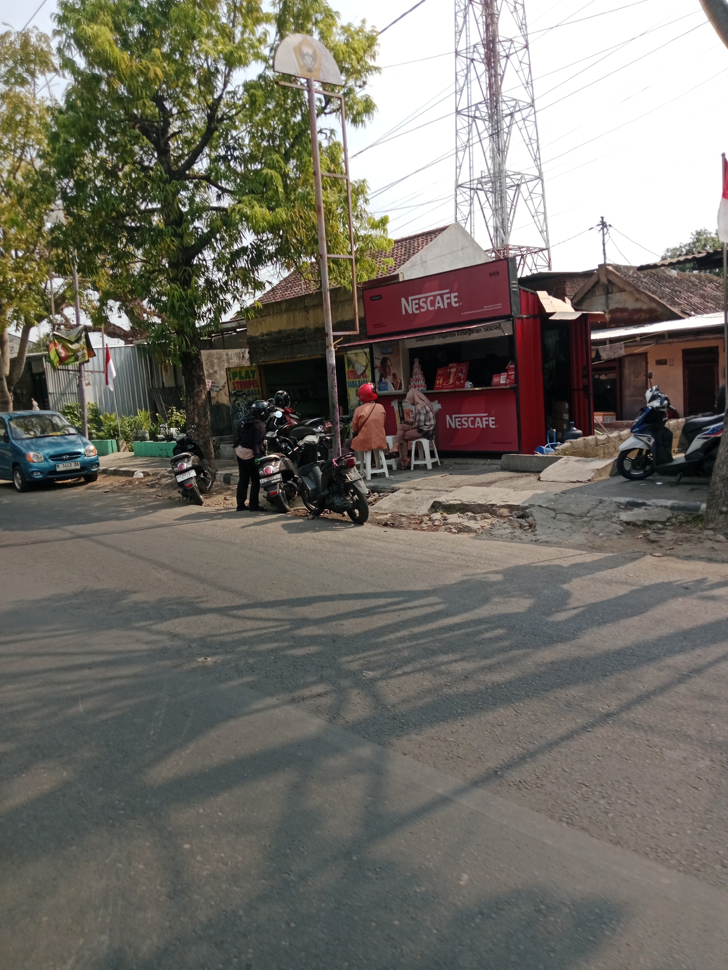 The image depicts a street scene in Kediri City, Indonesia, likely during daylight. A prominent red Nescafe kiosk is situated on the right side of the frame, featuring prominent branding and a service counter. Two individuals are seated on stools in front of the kiosk, seemingly interacting with a vendor or waiting for service. Several motorcycles are parked along the sidewalk and in front of the kiosk, indicating a common mode of transportation in the area. A blue car is parked on the left side of the street, near a tree. The background shows residential buildings and a tall telecommunications tower. The street is paved, and shadows of trees and possibly a basketball hoop pole are cast across the road, suggesting a sunny day.
