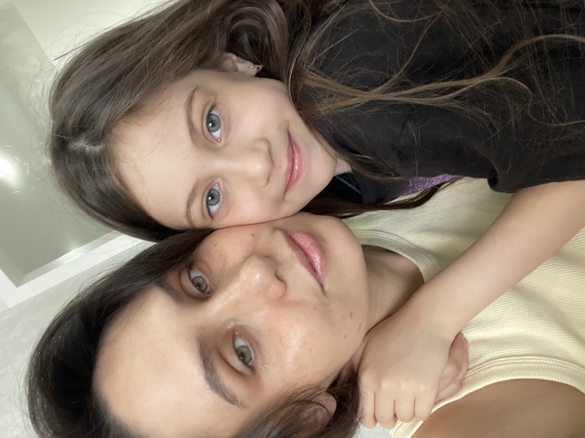 A selfie depicts a young girl with long brown hair and gray-blue eyes lying on top of a woman with dark hair and green eyes. The girl is wearing a dark-colored shirt, and the woman is wearing a light yellow shirt. The background is a blurred white wall. The location is not discernible from the image.