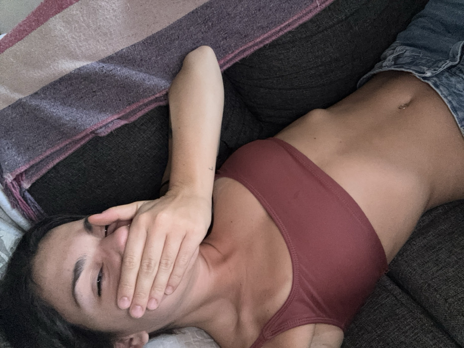 A woman lying on a gray couch, covered by a blanket. She wears a maroon-colored sports bra and blue denim shorts. Her hand covers her mouth, obscuring part of her face. The woman's head rests on a pillow. Her dark hair is pulled back. The photograph captures a relaxed and casual moment.