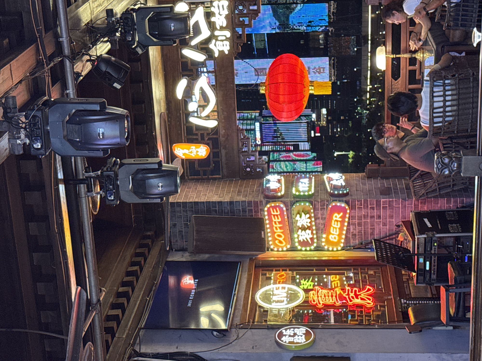 The image depicts an outdoor bar or restaurant setting at night, likely in the Yuzhong District of China, given the presence of Chinese neon signs and architectural elements. In the foreground, several people are seated at a table, engaged in conversation. The environment is illuminated by a mix of neon signs, overhead lights, and the city lights in the background.
Specific details include:
* **Location:** An outdoor seating area with a view of a city skyline at night. The presence of Chinese signage suggests a location within China.
* **People:** At least five individuals are visible, seated at a table. They appear to be interacting socially.
* **Objects:**
* **Neon Signs:** Numerous neon signs are present, indicating a commercial establishment. Some signs are in English, such as "LIVE," "COFFEE," "BEER," and "TEA," while others are in Chinese characters. A prominent red lantern hangs in the mid-ground.
* **Lighting:** Stage lighting fixtures are suspended from the ceiling, suggesting the venue might host live performances. A table lamp is on the table where the people are seated.
* **Furniture:** Wicker chairs and tables are arranged in the seating area. In the foreground, there is a music setup with speakers, a mixer, and a music stand.
* **Architecture:** Wooden beams and decorative elements are visible in the ceiling and the structure of the building.
* **Environment:** The scene is dimly lit, with the neon signs providing vibrant colors. The background shows a dense urban cityscape with illuminated buildings and lights. The overall atmosphere is lively and convivial.
* **Actions:** People are seated, talking, and potentially dining or drinking. The presence of the music equipment suggests that live entertainment may be part of the establishment's offerings.