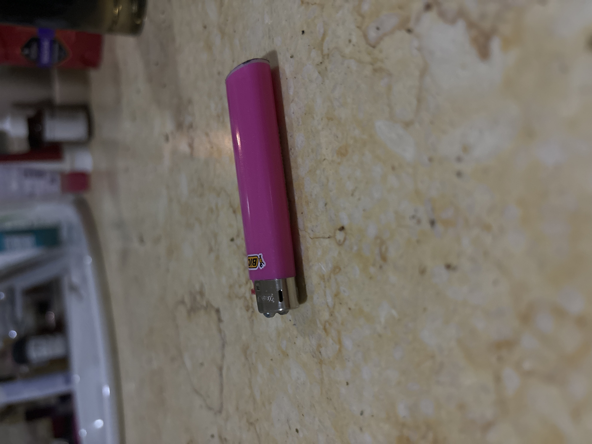 A pink BIC lighter rests on a surface with a beige marbled pattern. A shelf of various items is visible in the background. The scene appears to be indoors. There is no indication of the city of Budva, Montenegro.