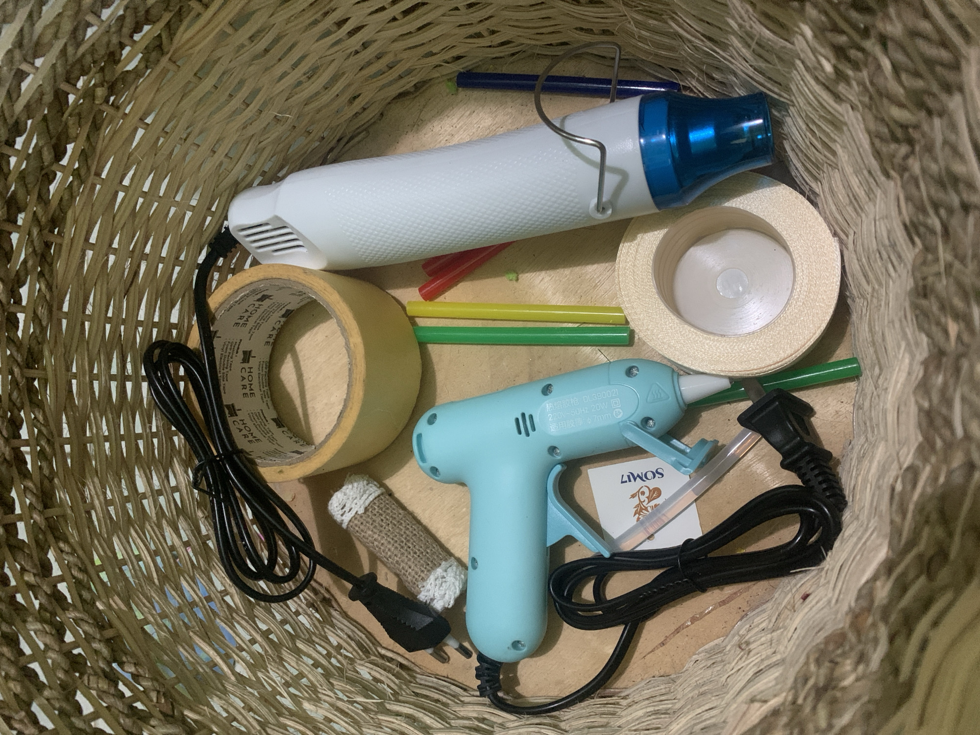 This image is an overhead shot of a woven basket filled with craft supplies. The basket is made of natural fibers and appears to be placed on a wooden surface. Inside the basket, there is a light blue glue gun with a black cord, a white hot glue stick, a roll of tan-colored tape, a white hot glue gun with a blue nozzle, several colorful glue sticks (green, yellow, red), a blue marker or pen, and a roll of light beige ribbon. There is also a small piece of burlap fabric with white yarn wrapped around it. The items are arranged somewhat haphazardly within the basket. The lighting is bright, suggesting an indoor environment. There are no people or animals present in the image. The scene depicts craft supplies ready for use. The question about the location in Kuala Pembuang, Indonesia is not supported by any visual evidence in the image.