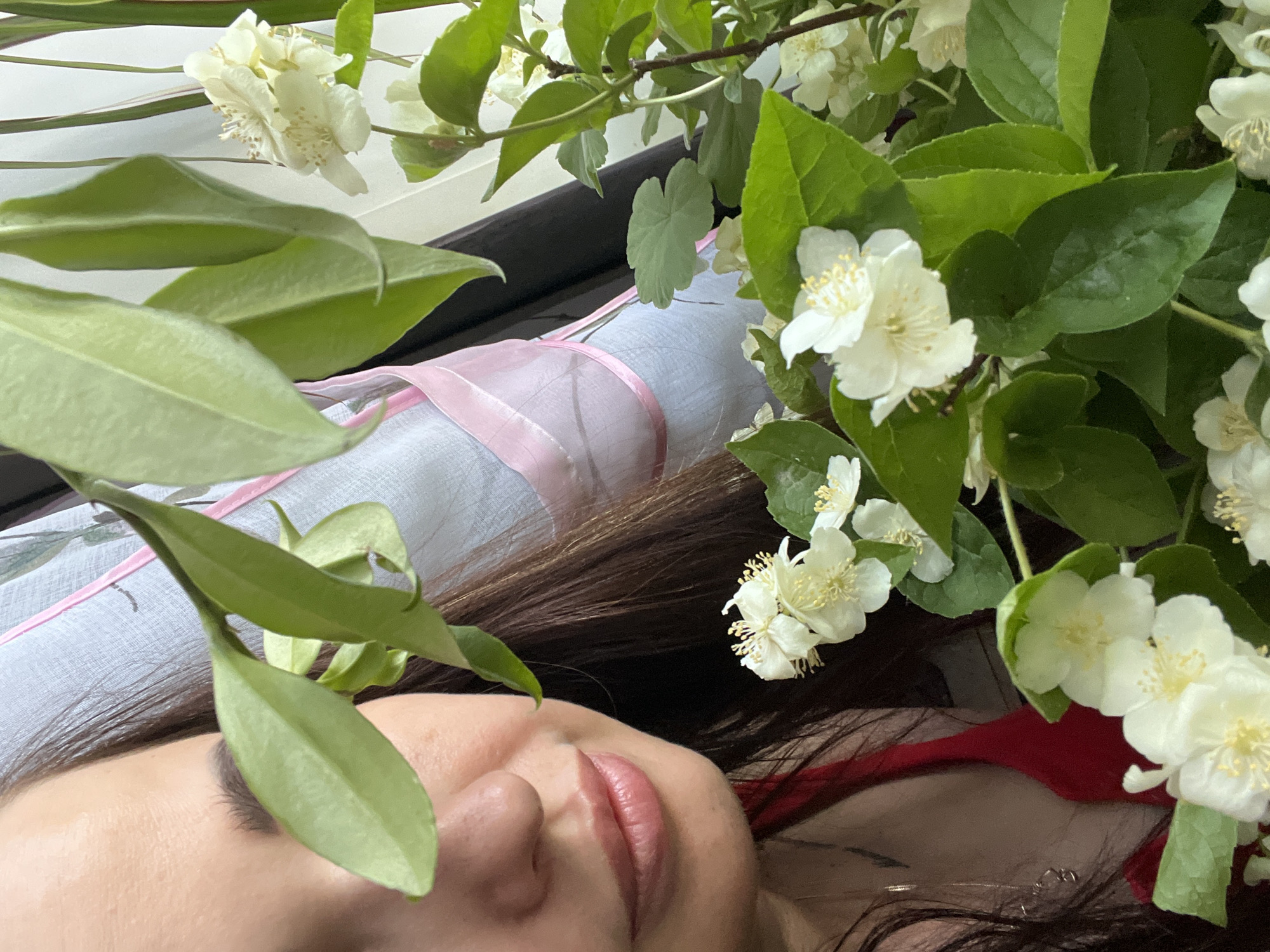 A close-up medium shot shows the left side of a woman's face, partially obscured by green leaves and white jasmine flowers. Her eye is visible, and the bridge of her nose and lips are also in focus. She is wearing a red sleeveless top. The background consists of a light-colored fabric with a pink ribbon, and dark curtains. The scene is indoors and appears to be a still life composition.