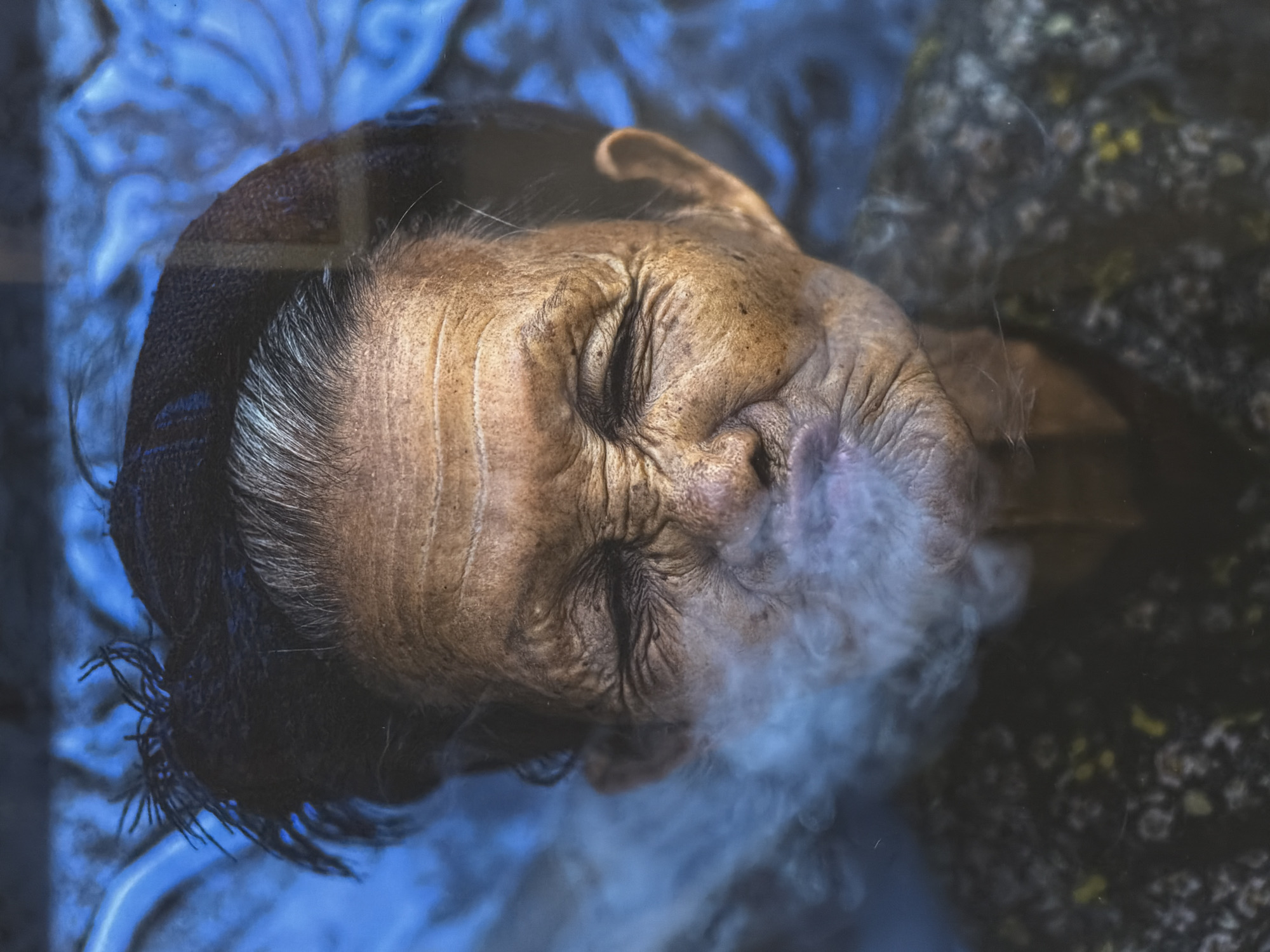 This is a close-up, high-angle portrait of an elderly person, likely a woman, captured in Hội An Ward, Vietnam. The subject's face is highly detailed, marked by deep wrinkles and age spots, and their eyes are closed, conveying a sense of serenity or concentration. Silver-gray hair is visible at the temples, contrasting with the warm, weathered skin tone. A plume of white smoke or vapor is exhaling from their mouth, dissipating into the lower right of the frame, suggesting an act of smoking or vaping.
The subject wears a dark head covering or garment that frames their face and neck. On the left side of the frame, behind the person's head, is a vibrant blue background with intricate, flowing patterns, possibly a textile or decorative wall. To the right, another patterned fabric is visible, dark with subtle, lighter floral or abstract designs, suggesting clothing or upholstery. The lighting emphasizes the texture and lines of the subject's face and the ethereal quality of the smoke, creating a contemplative atmosphere. There is no visible text.