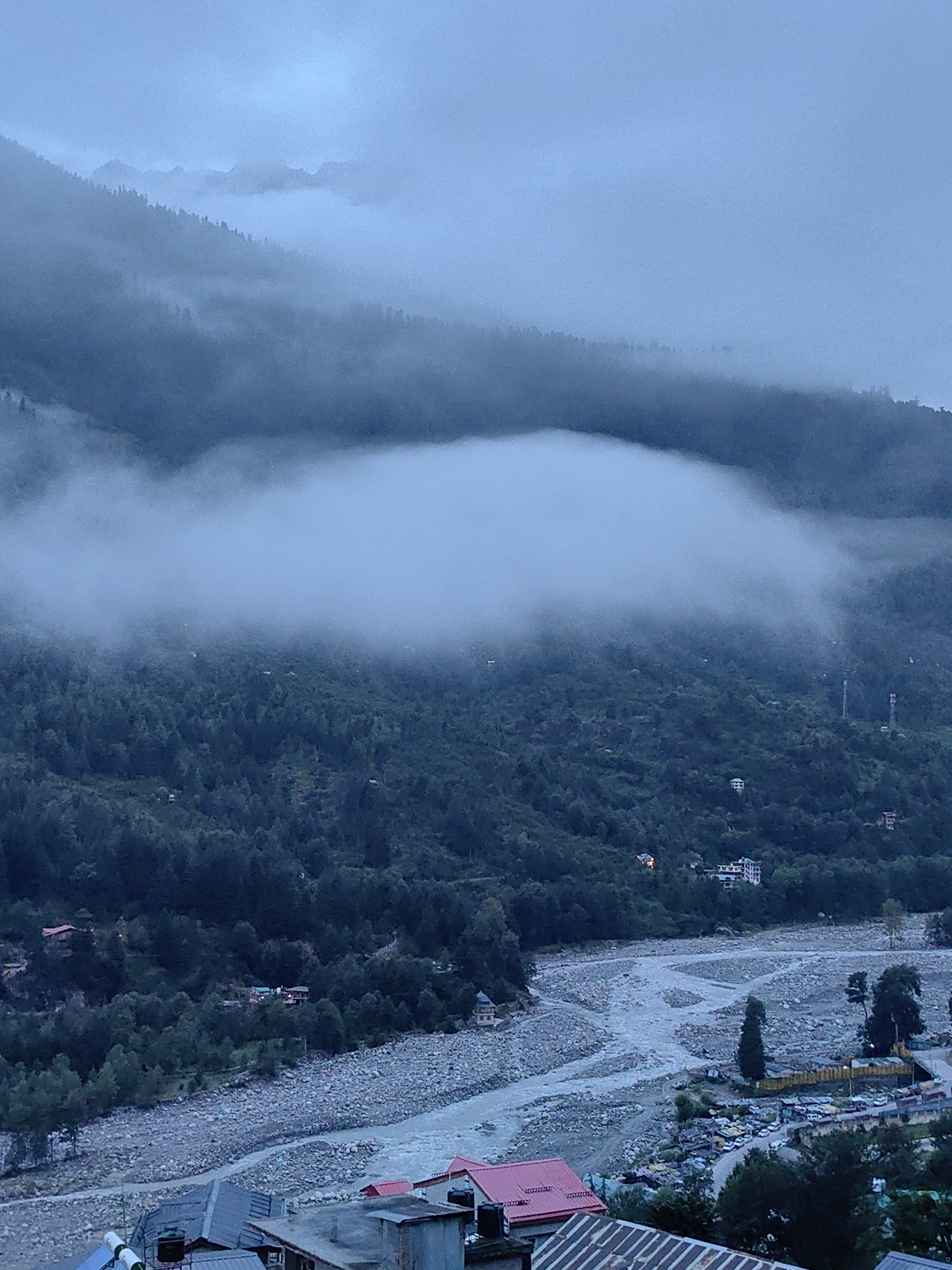 The image depicts a landscape in Vashisht, India, under a cloudy and misty sky. The foreground features a cluster of buildings with various roof types, including red and grey corrugated metal. Adjacent to these structures is a wide riverbed filled with stones, through which a portion of the river flows. On the far bank of the river, a road is visible, lined with parked cars and some buildings.
Further into the scene, a densely forested mountainside rises, with trees creating a dark green expanse. A large mass of white clouds hangs low, partially obscuring the upper slopes of the mountain and creating a hazy, atmospheric effect. The mountain peaks in the distance are barely visible through the mist and clouds, contributing to a subdued and muted color palette dominated by blues and greys. The overall impression is one of a serene, somewhat overcast day in a mountainous region.
