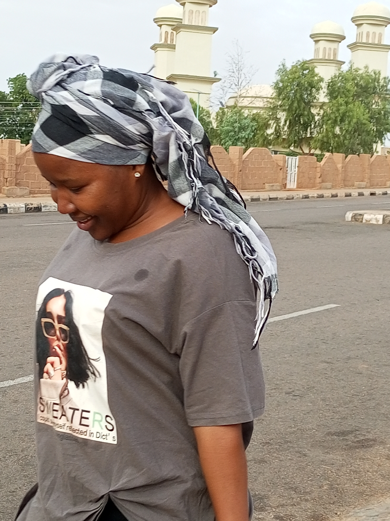 A young Black woman wearing a grey t-shirt with a graphic print and a black, white, and grey patterned headwrap stands outdoors in front of a building with domes and arches, possibly a mosque or government building. The woman is smiling and looking down to her left. Her t-shirt has a graphic of a woman wearing sunglasses and the text "SWEATERS" and "could see yourself reflected in Dict's" printed on it. She is wearing a small diamond stud earring. The background includes a brick wall and a paved road. The scene appears to be in the city of Gombe, Nigeria.
