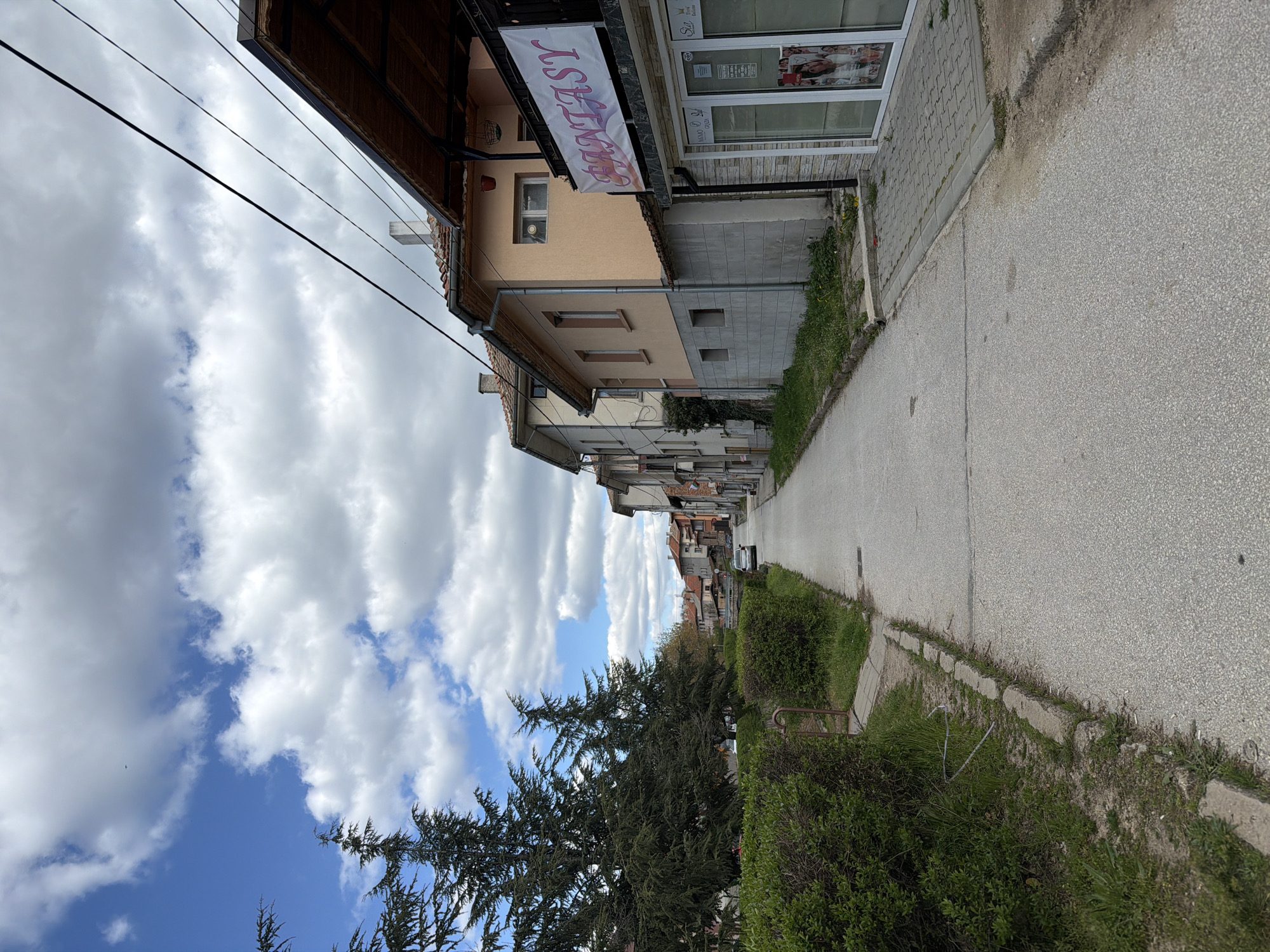 This media file depicts a street scene in Panagyurishte, Bulgaria, under a partially cloudy sky. The image shows a narrow, paved road or pathway extending into the distance, with buildings lining one side and vegetation on the other.
On the left side, a row of buildings features diverse architecture. The nearest structure has a pale yellow-orange facade with a brown tiled roof and a prominent white banner displaying the word "FANTASY" in stylized pink and purple lettering. Adjacent to it, a shopfront with large display windows is visible, featuring posters and partially discernable text like "SZP" and "Luxury." This building transitions to a gray concrete base with grass and weeds growing along its foundation. Further down the street, more buildings with tiled roofs and various colors line the route. Black utility wires stretch across the upper portion of the sky.
On the right side of the road, a lush green area is present, dominated by a large, dark green coniferous tree with dense needles. Smaller bushes and shrubs also dot this side, along with patches of grass and some concrete elements, including curbing. The sky above is a mix of bright blue and fluffy white cumulus clouds, suggesting pleasant daytime weather, likely late morning or afternoon. No people or active events are depicted, lending the scene a quiet, everyday ambiance.