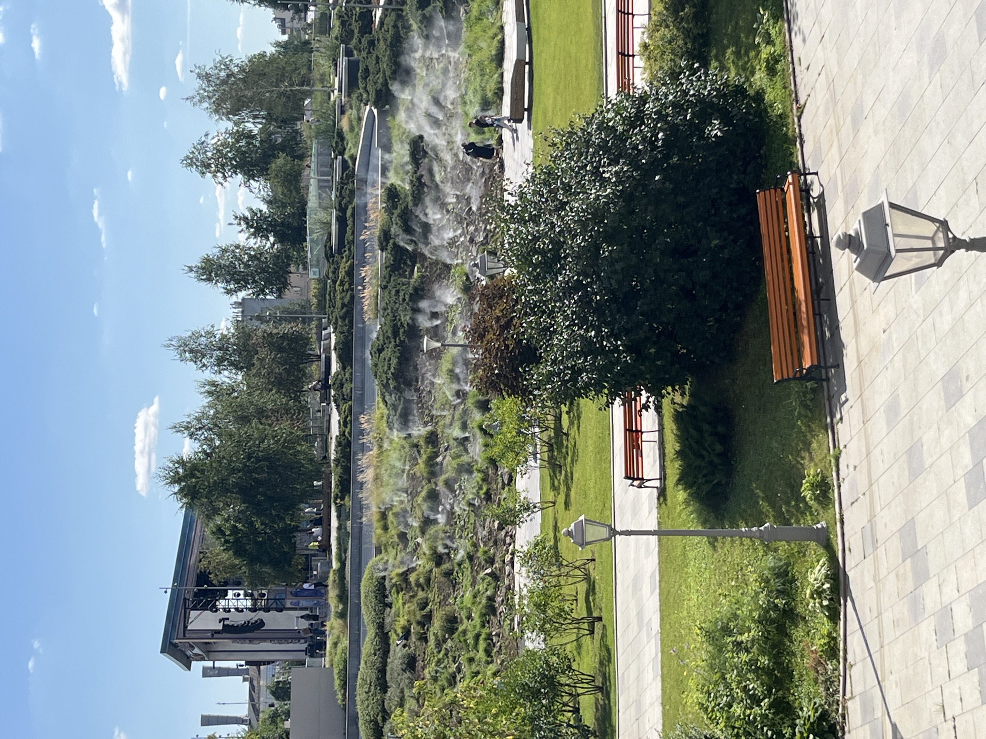 The image depicts a park with a contemporary design, featuring tiered landscaping and misting systems creating a hazy effect on the vegetation. In the background, a modern building with a large stage structure is visible, suggesting an entertainment venue. Several trees are scattered throughout the park, some in the foreground and others forming a backdrop.
The foreground shows a paved walkway with a distinctive patterned tile design. A vintage-style street lamp stands to the left of the walkway, casting a shadow. Two orange benches are placed on the grassy area adjacent to the walkway. A large, dark green bush dominates the mid-right portion of the frame.
Further back, the tiered landscape is covered in lush green grass and shrubs, with mist emanating from numerous emitters, creating a diffused atmosphere. People can be seen walking and sitting in this area, some interacting and others appearing to be enjoying the park. The sky is a clear blue with a few scattered white clouds.
The overall scene suggests a public park in Moscow, Russia, designed for leisure and possibly outdoor events, given the presence of the stage. The misting system adds a unique aesthetic to the landscape.