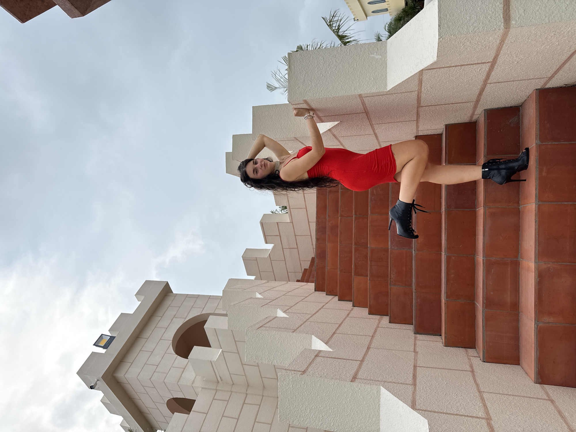 A young woman with long, dark hair, dressed in a vibrant red form-fitting mini-dress and black high-heeled ankle boots, poses for a photograph. She leans against a textured white block structure, with her arms raised behind her head, and looks towards the camera with a gentle smile. A silver bracelet adorns her right wrist.
The setting features an ornate staircase with terracotta-colored tiled risers and treads, bordered by striking white, block-like architectural elements that create a castle-like aesthetic. To the woman's left, a light-colored building with arched windows, block patterns, and crenellated top edges reinforces this theme. A floodlight and a small security camera are visible on this building.
The background consists of an overcast sky, suggesting a cloudy daytime. Sparse green foliage, possibly palm leaves, and the upper part of another light-colored building can be seen in the distance. The overall scene conveys a playful and confident mood amidst an elaborately designed outdoor space, likely a resort area in Mui Ne, Vietnam. There is no visible text in the image.